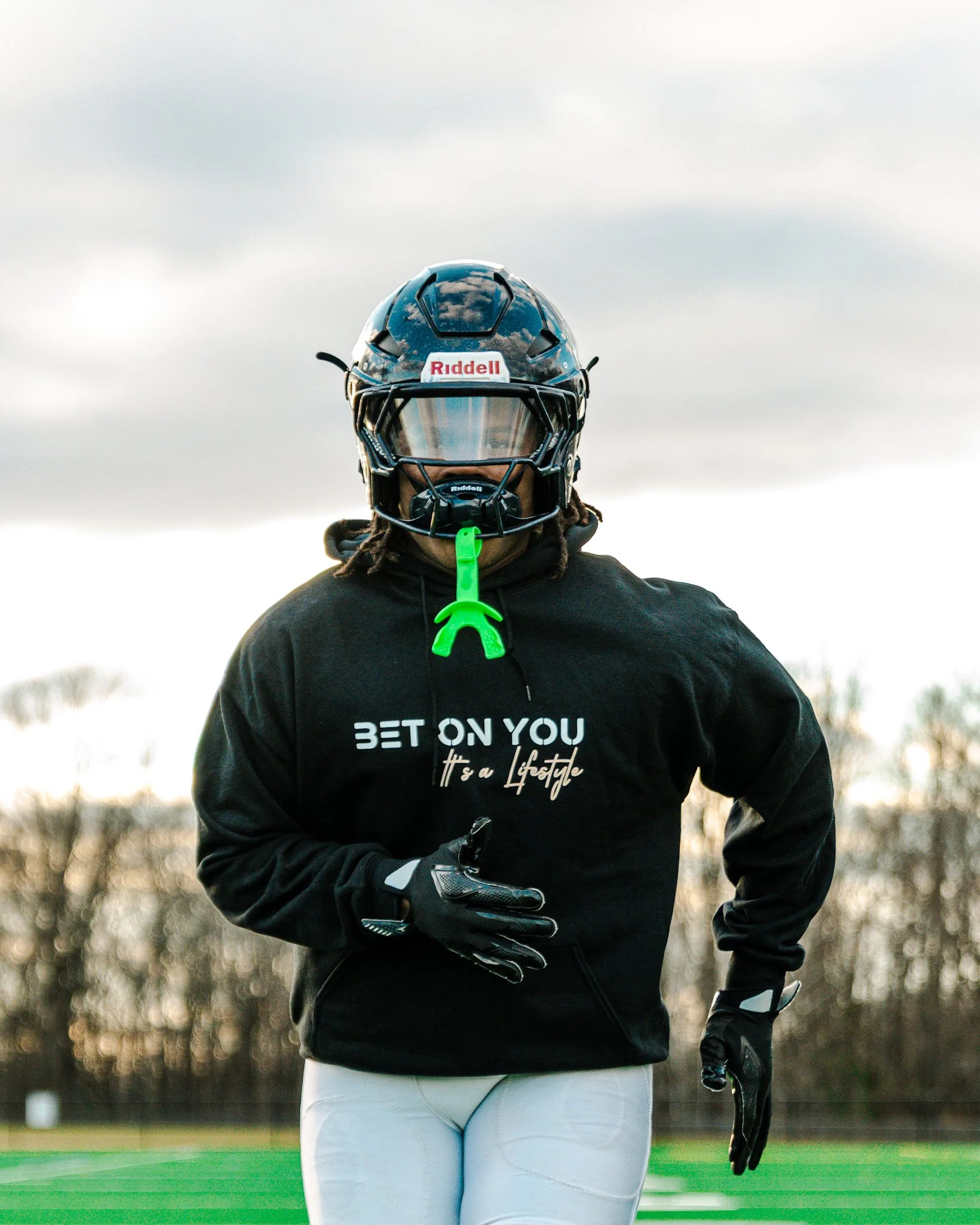 A football player standing on a field wearing a black Riddell helmet, black hoodie with yellow and white text, and white football pants, with trees and a cloudy sky in the background.