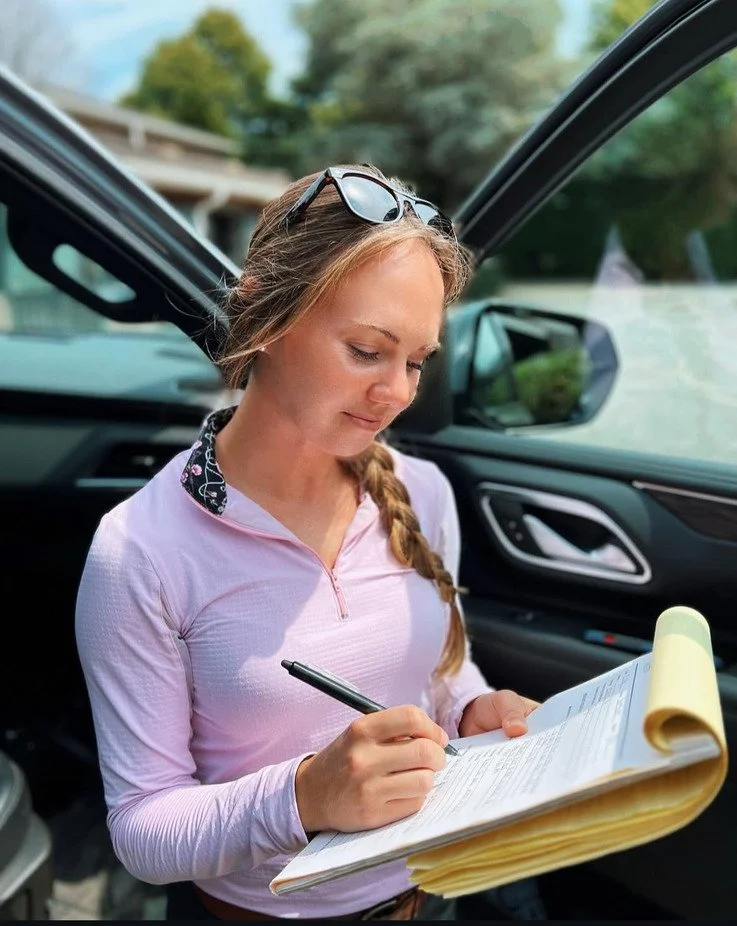 A woman with sunglasses on her head, wearing a light pink long-sleeve top, is standing next to an open car door, writing on a large clipboard or notepad with a black pen, outdoors.