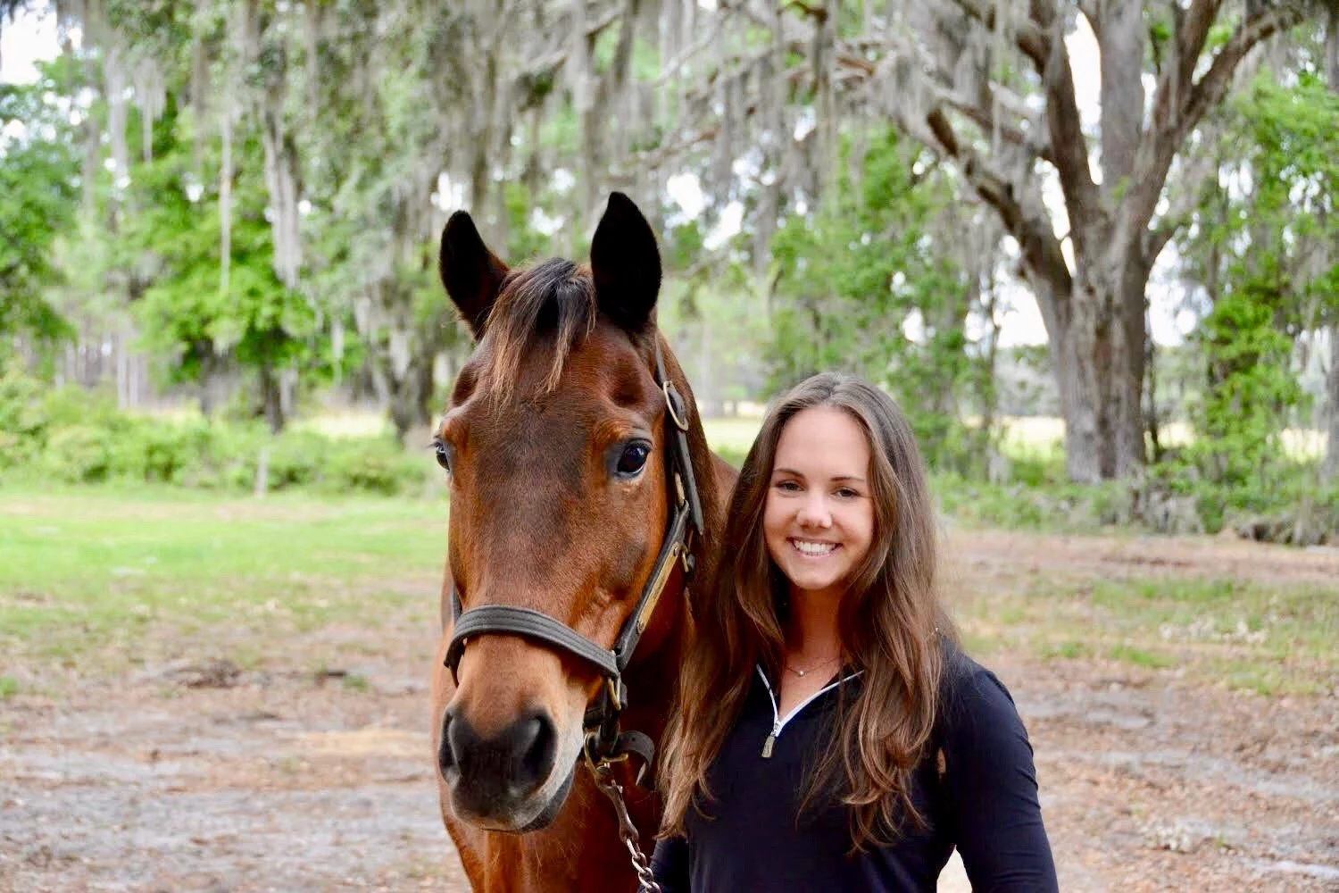 A woman with long brown hair smiling next to a brown horse with a black mane in an outdoor setting with trees and grass in the background.