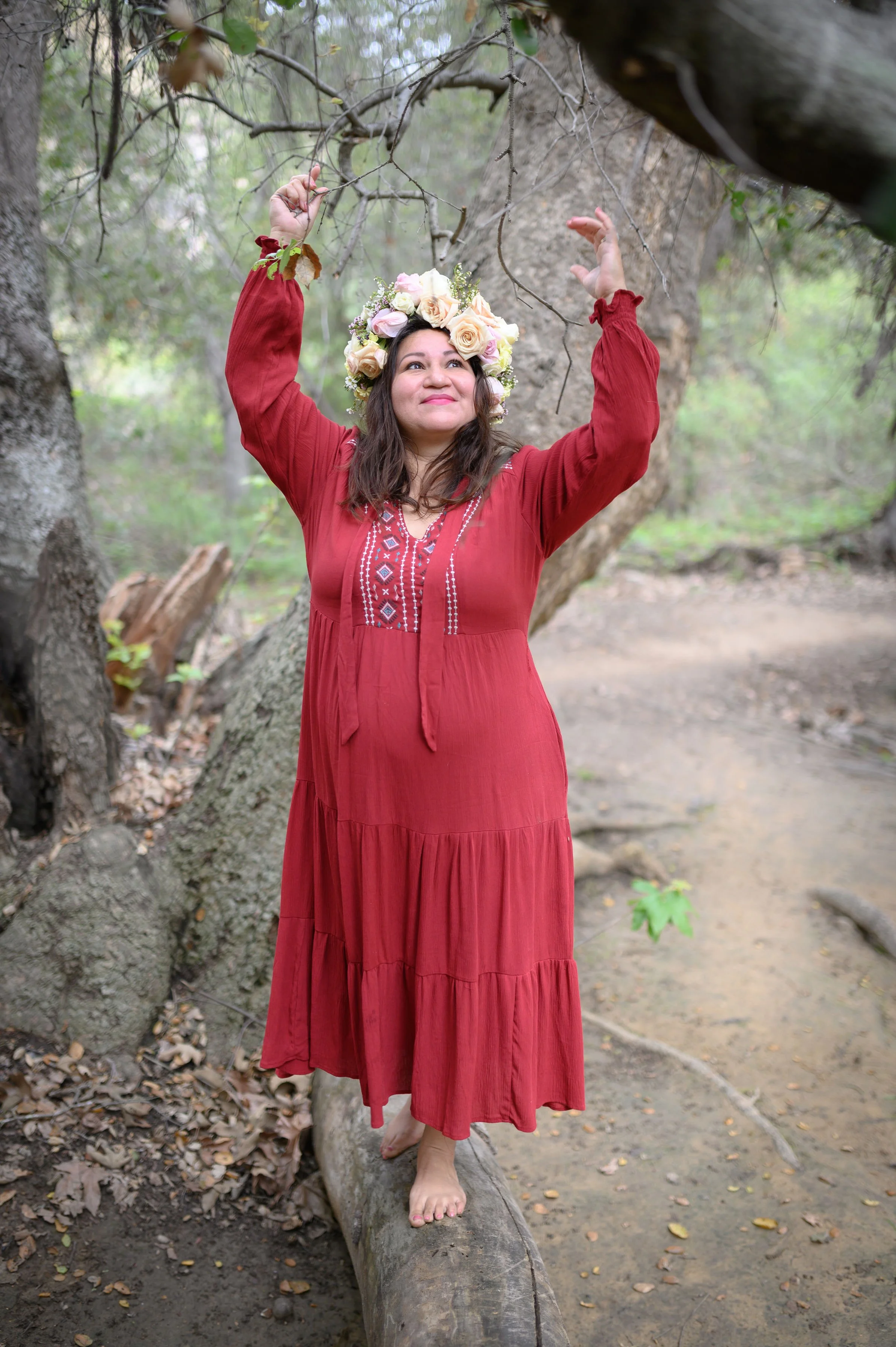 A woman in a red dress with embroidered details and a flower crown stands barefoot on a fallen log in a forest, reaching up to touch the tree branches overhead.