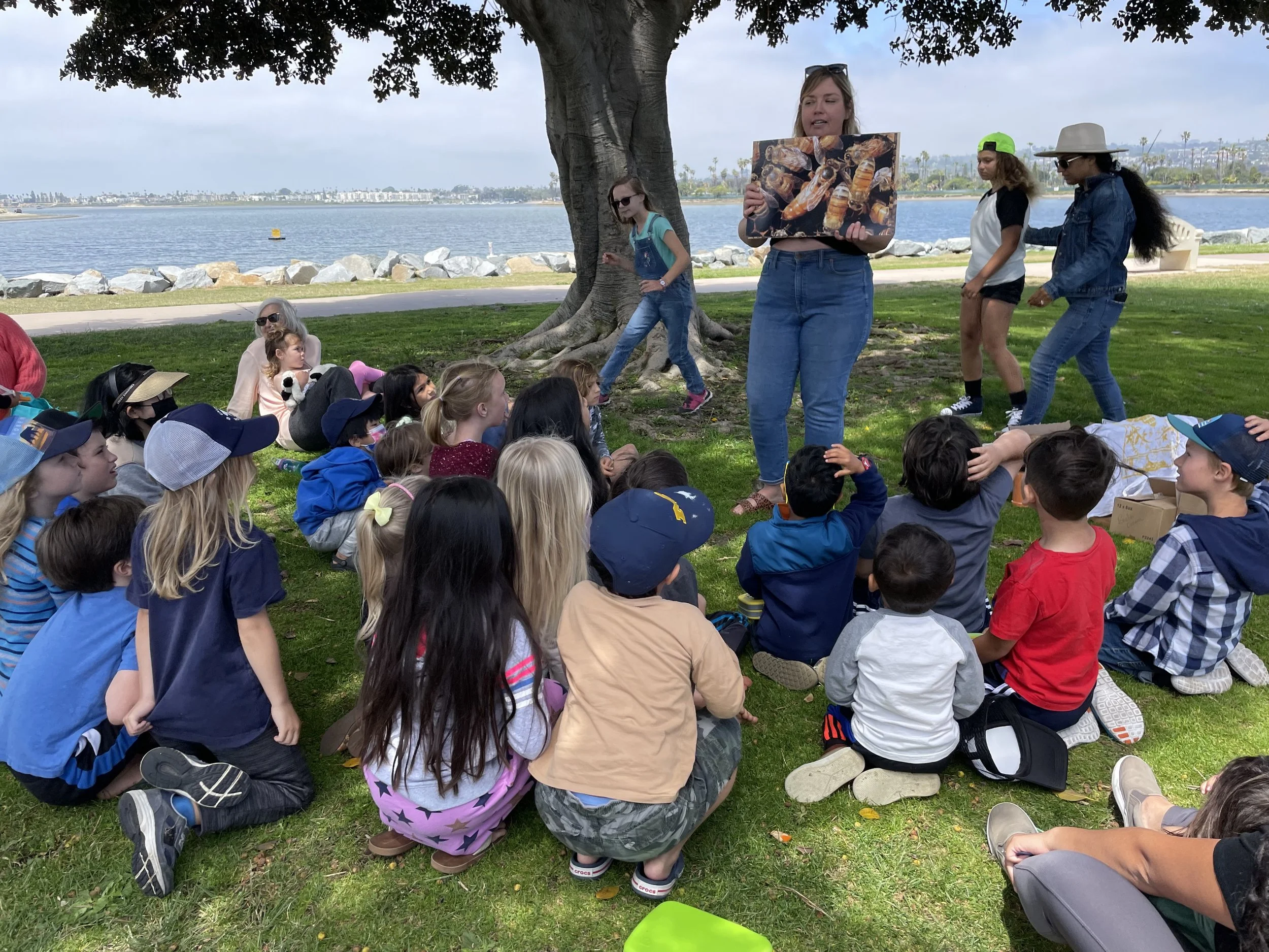 A woman stands under a tree, holding a large picture of bees, addressing a group of children seated on the grass near a waterfront. Others walk around or sit on the grass, with some adults nearby.