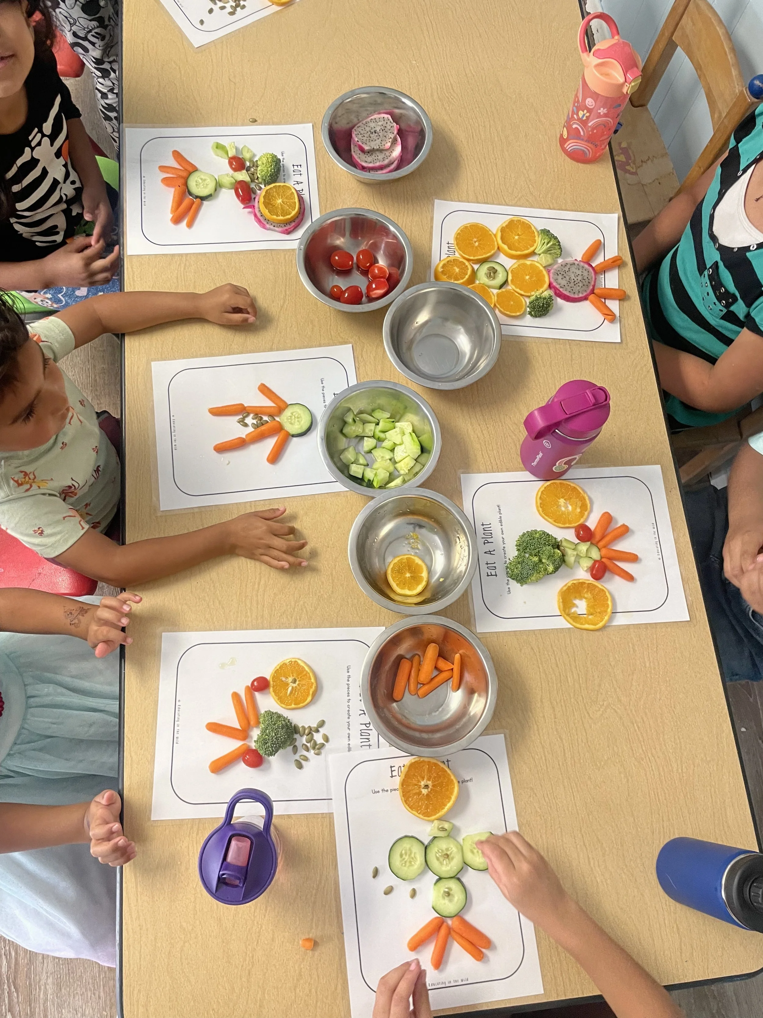 Children arranging fresh vegetables and fruits on paper placemats, including cucumber, carrots, cherry tomatoes, broccoli, orange slices, and dragon fruit, on a wooden table.