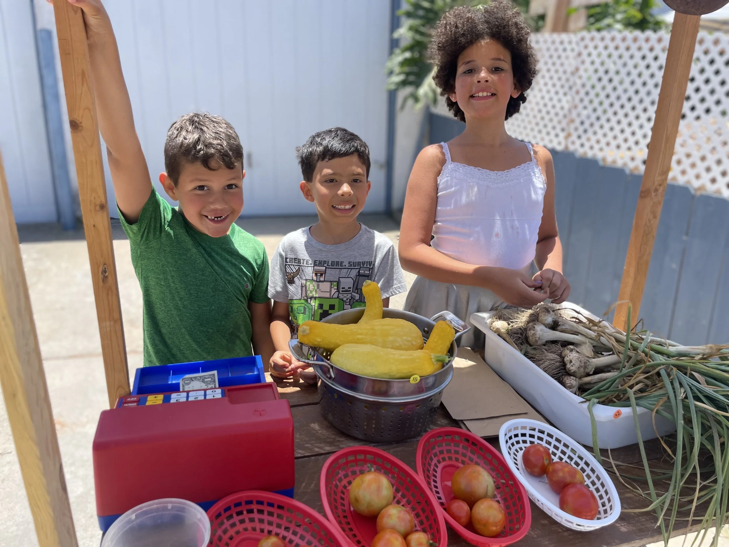 Three children at a vegetable stand outdoors with fresh produce including squash, garlic, and tomatoes, smiling and looking at the camera.