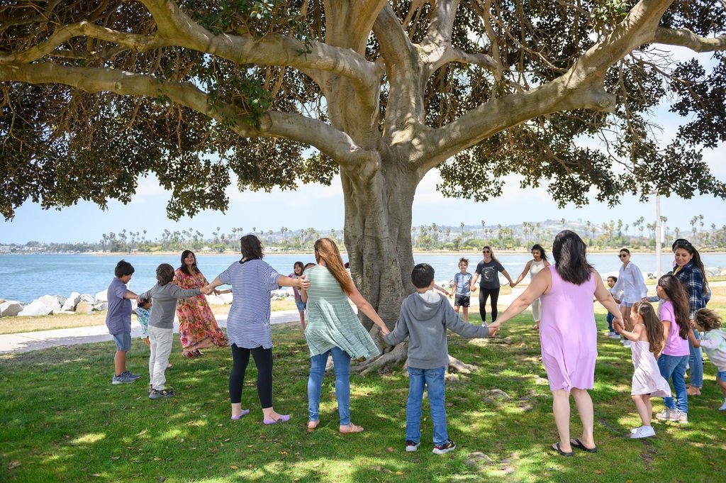 Group of children and adults holding hands, forming a circle under a large tree near a body of water, enjoying outdoor activity.