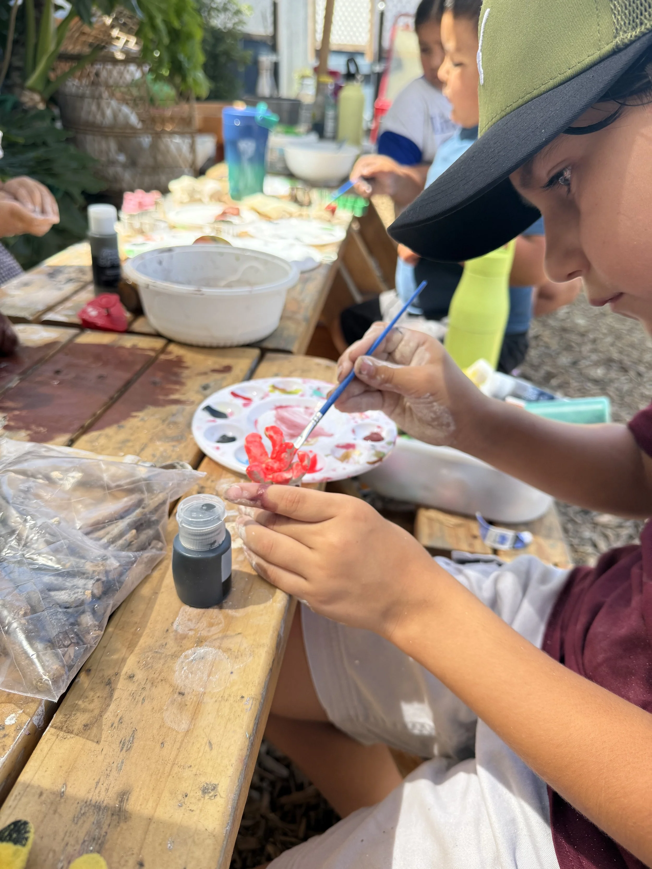 Children painting with red and black paint at an outdoor art table.