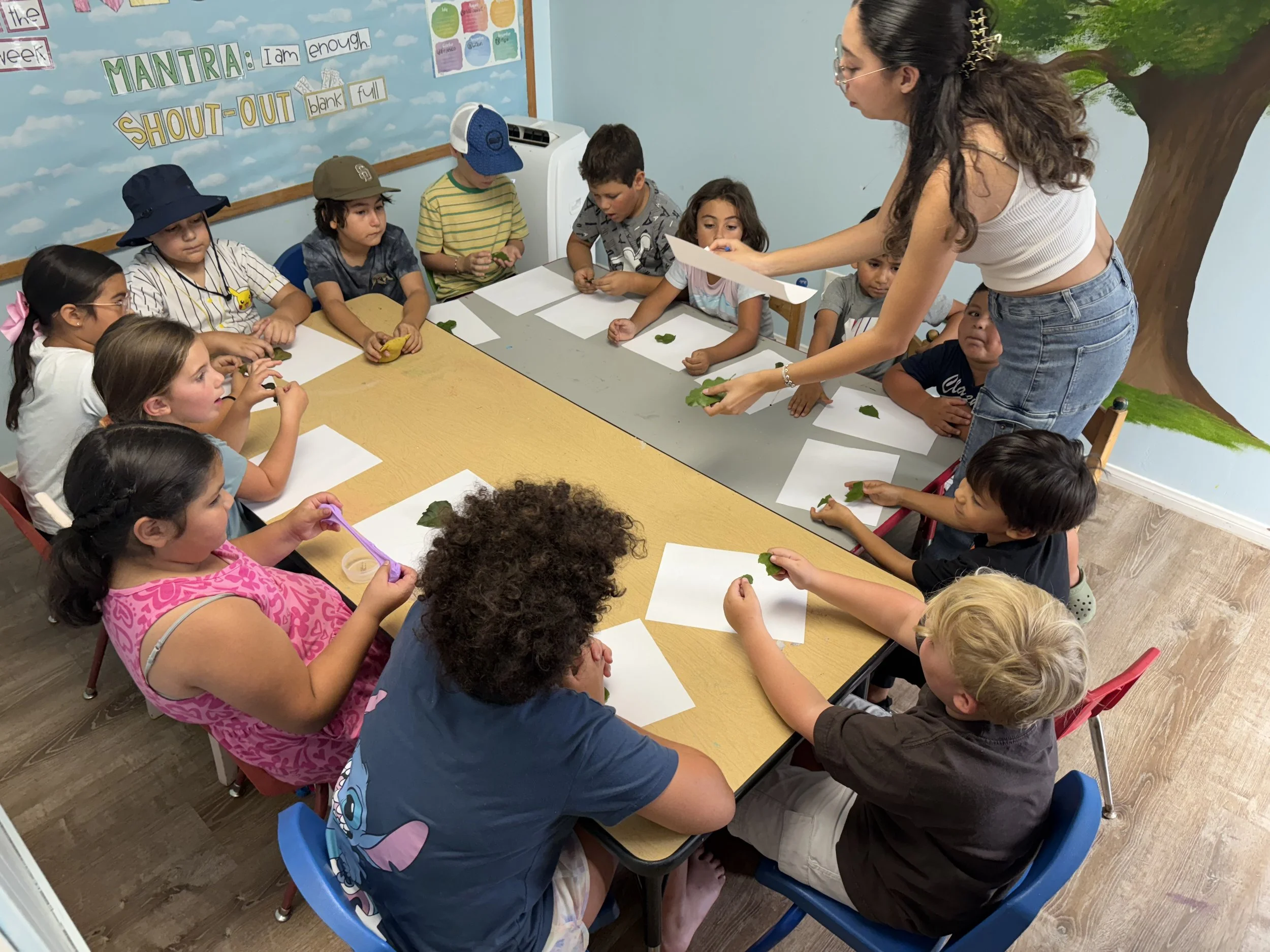 A teacher hands out green leaves to children sitting at a table in a classroom, with a blue wall featuring a tree mural in the background.