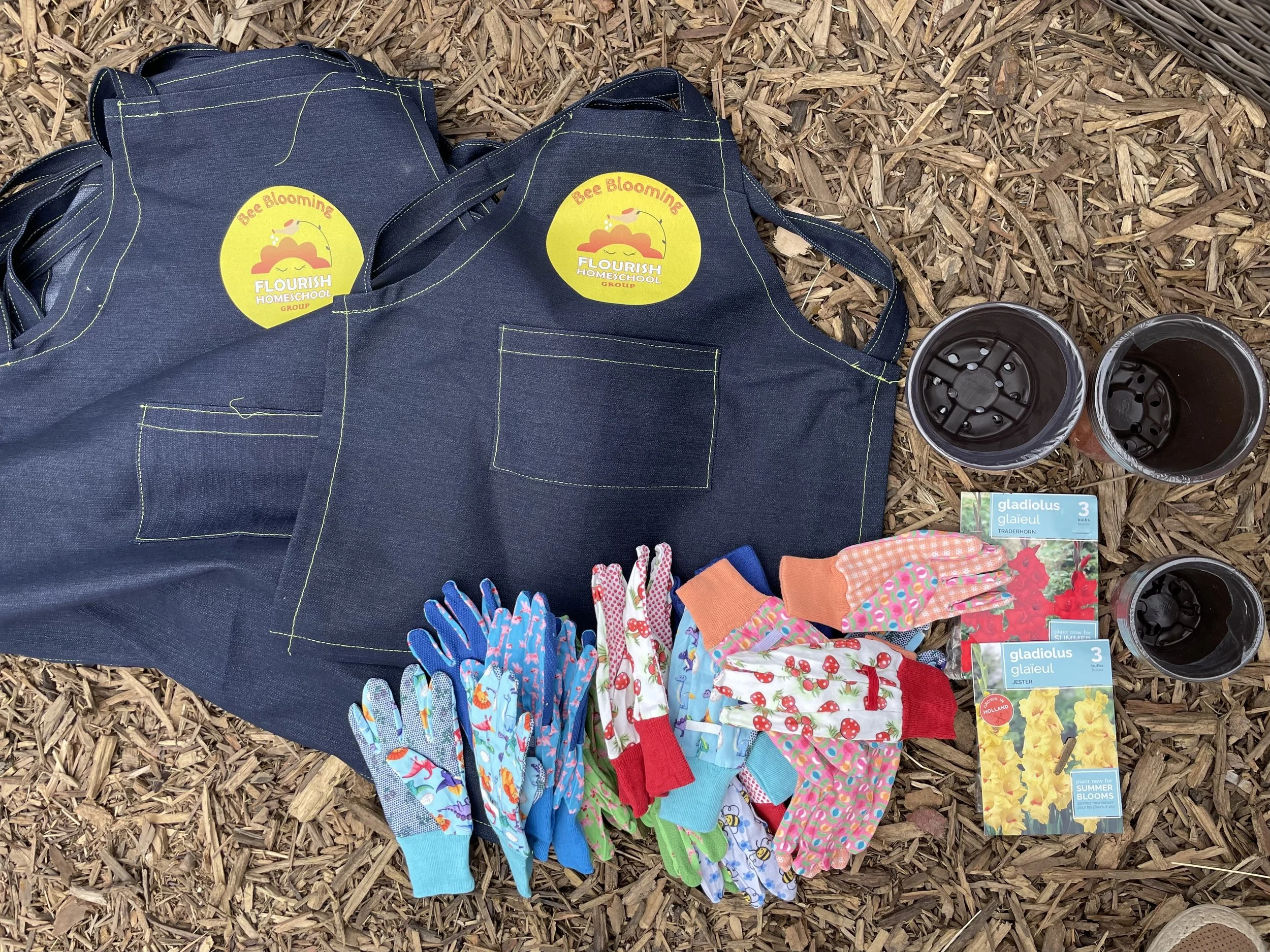 Gardening supplies including a black denim apron with a yellow "Bee Blooming" sticker, multiple pairs of colorful gloves, three small black pots, and seed packets for gladiolus flowers on a bed of wood chips.