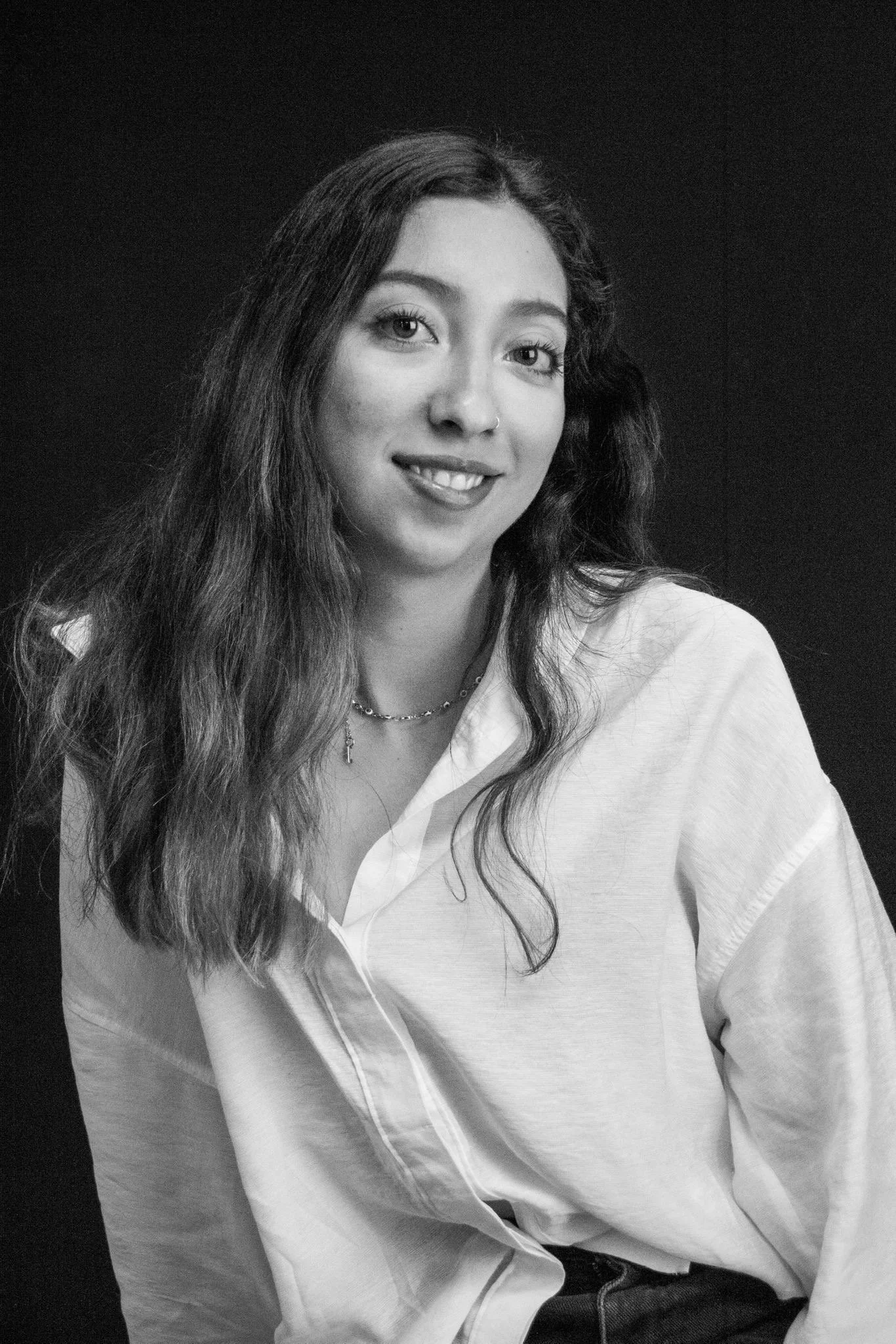 Portrait of a young woman with long wavy hair, smiling, wearing a light-colored shirt and a necklace, against a dark background.