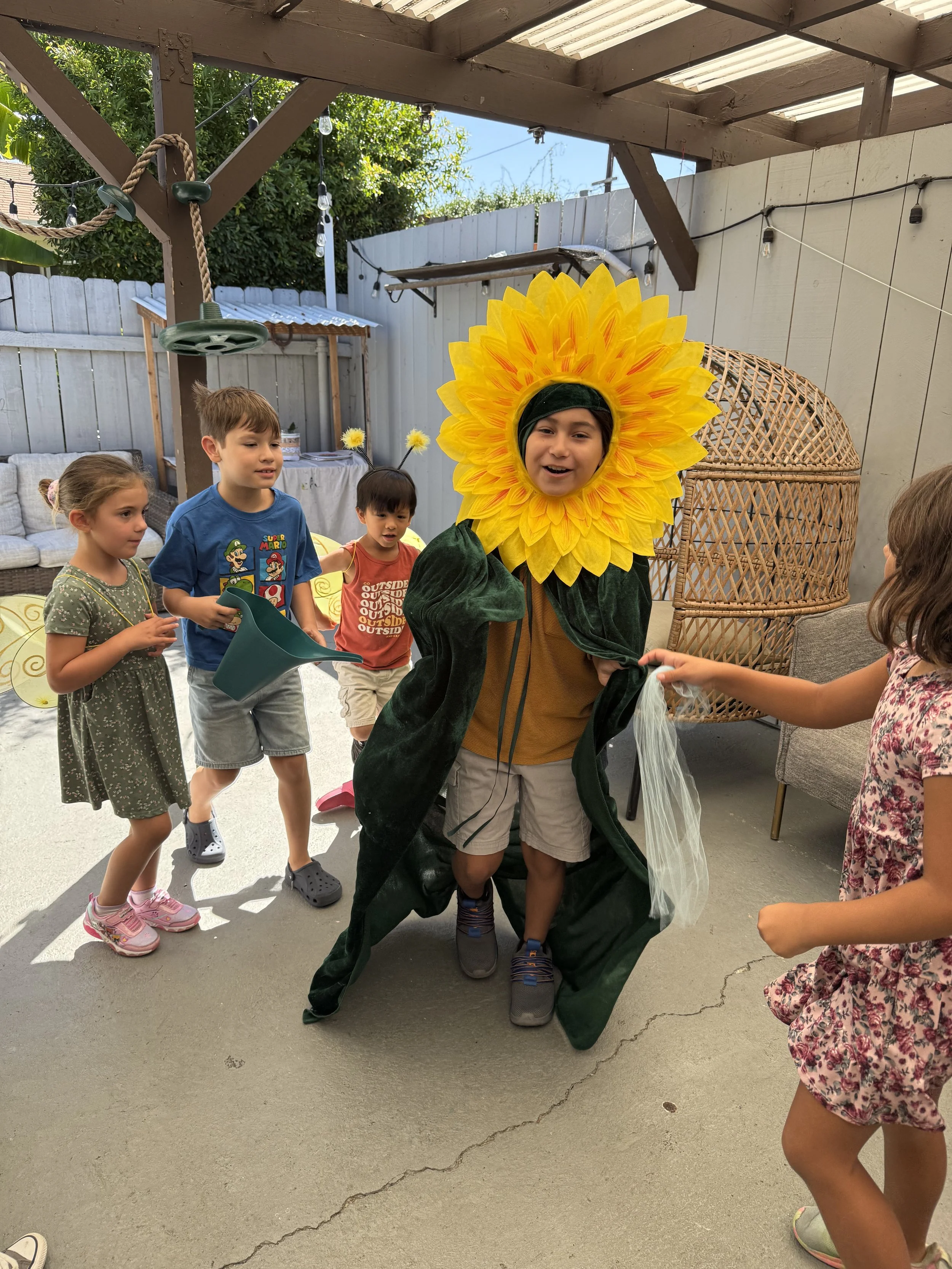 Child dressed as a sunflower with a large yellow petal headpiece, surrounded by children on a porch or backyard area.