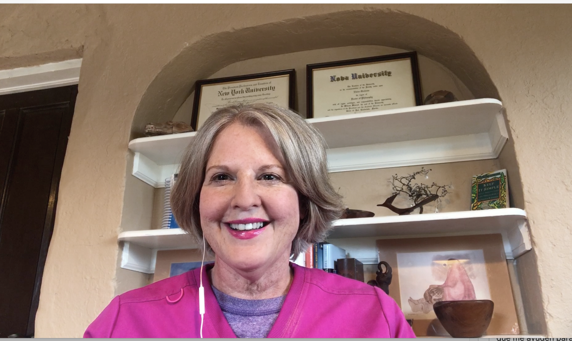 A photo of Dr. Elaine Foster, PhD at her desk wearing scrubs and white earbuds, standing in front of a white shelf with framed diplomas from New York University, decorative items, and books.