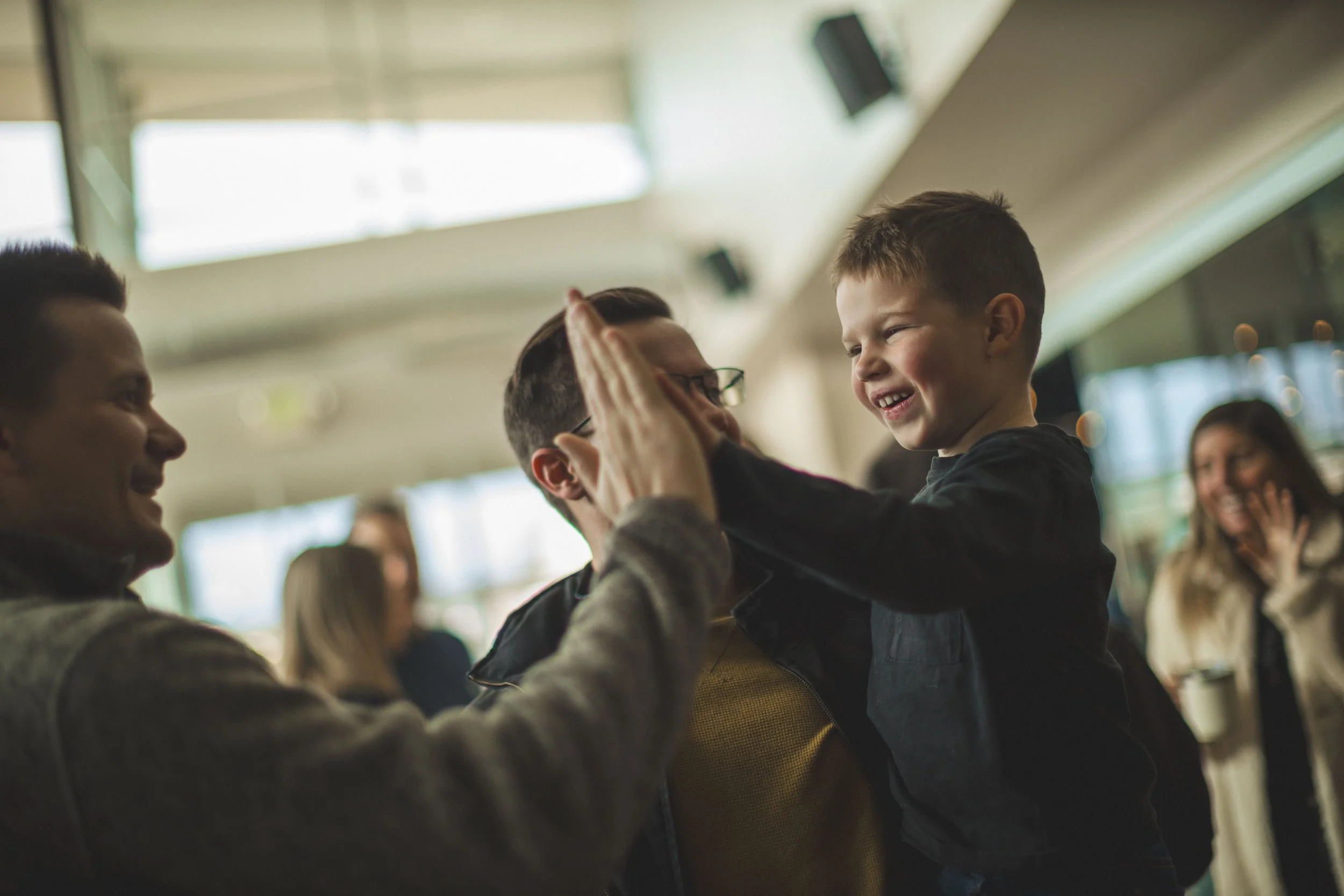 A young boy and two men smiling indoors, with one man and the boy giving each other a high five.