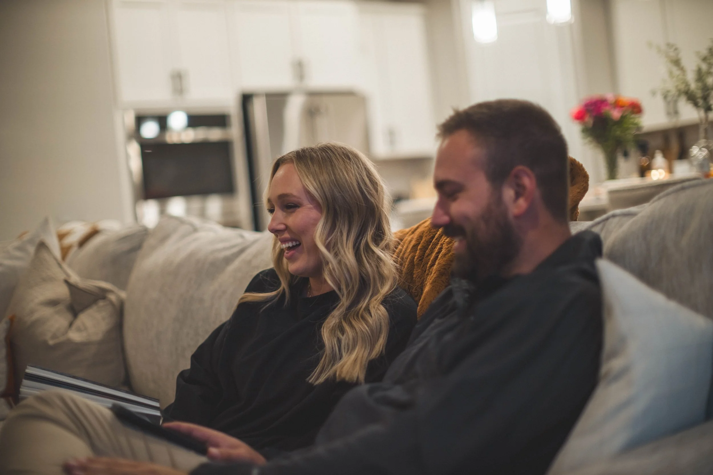 A woman and man sitting on a beige couch, smiling and looking at a tablet together in a living room with a kitchen in the background.
