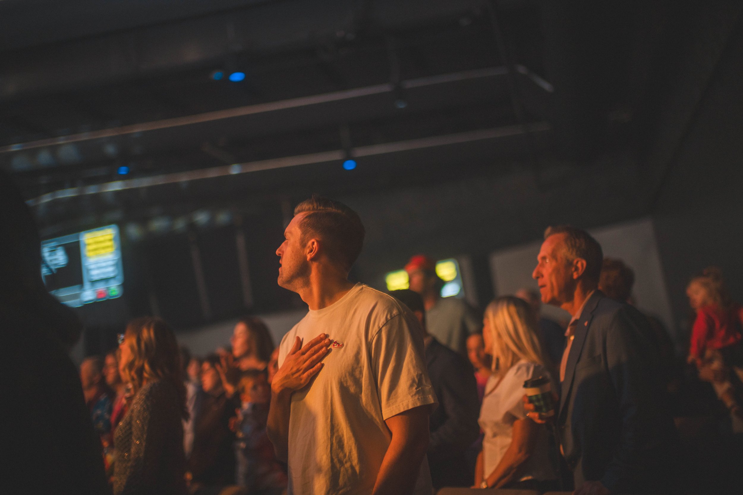 People standing in a darkened room, some with hand over heart, appearing to be engaged in a moment of silence or prayer.