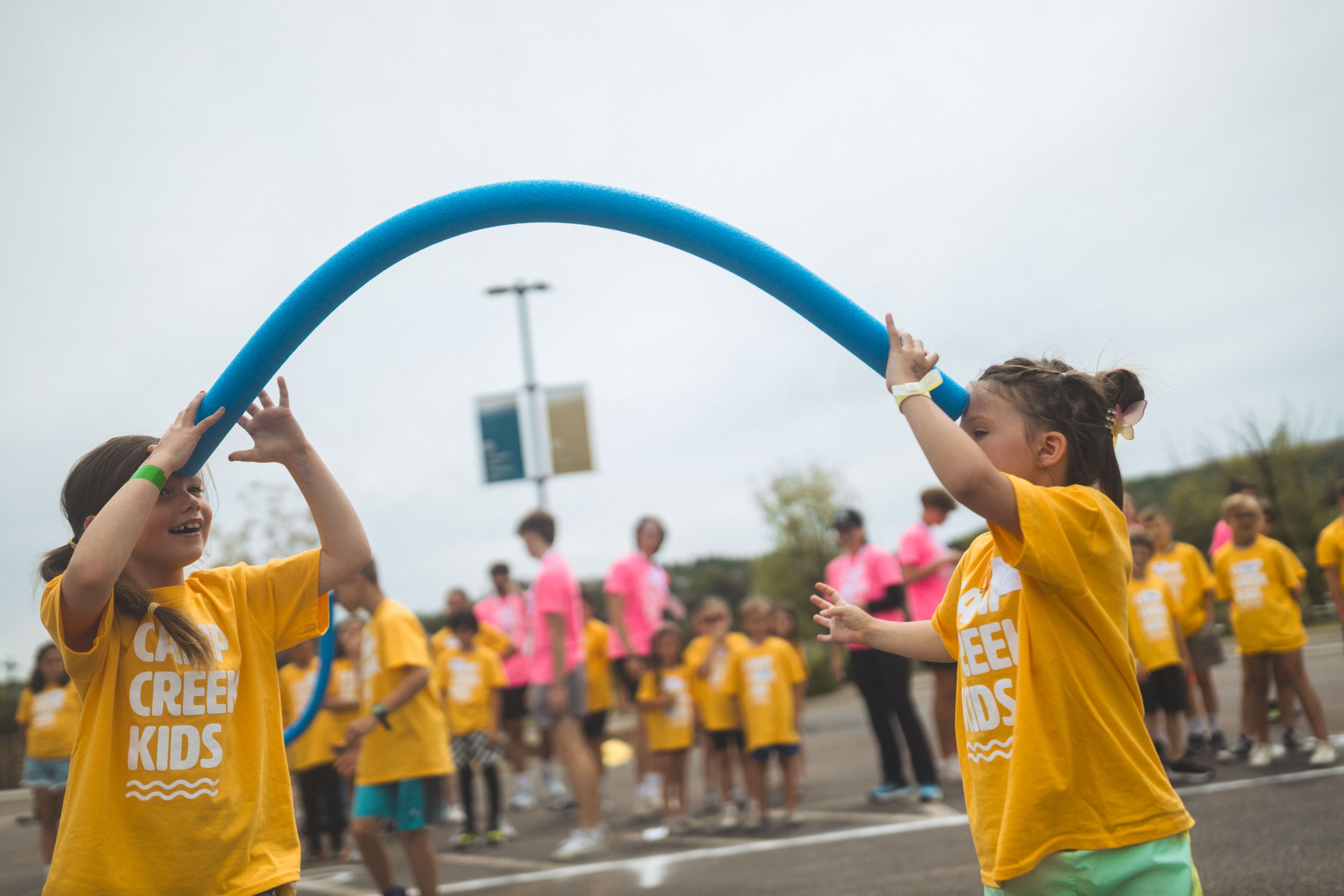 Two young girls playing with a blue foam noodle during a kids' camp event outdoors, with children and adults in the background.