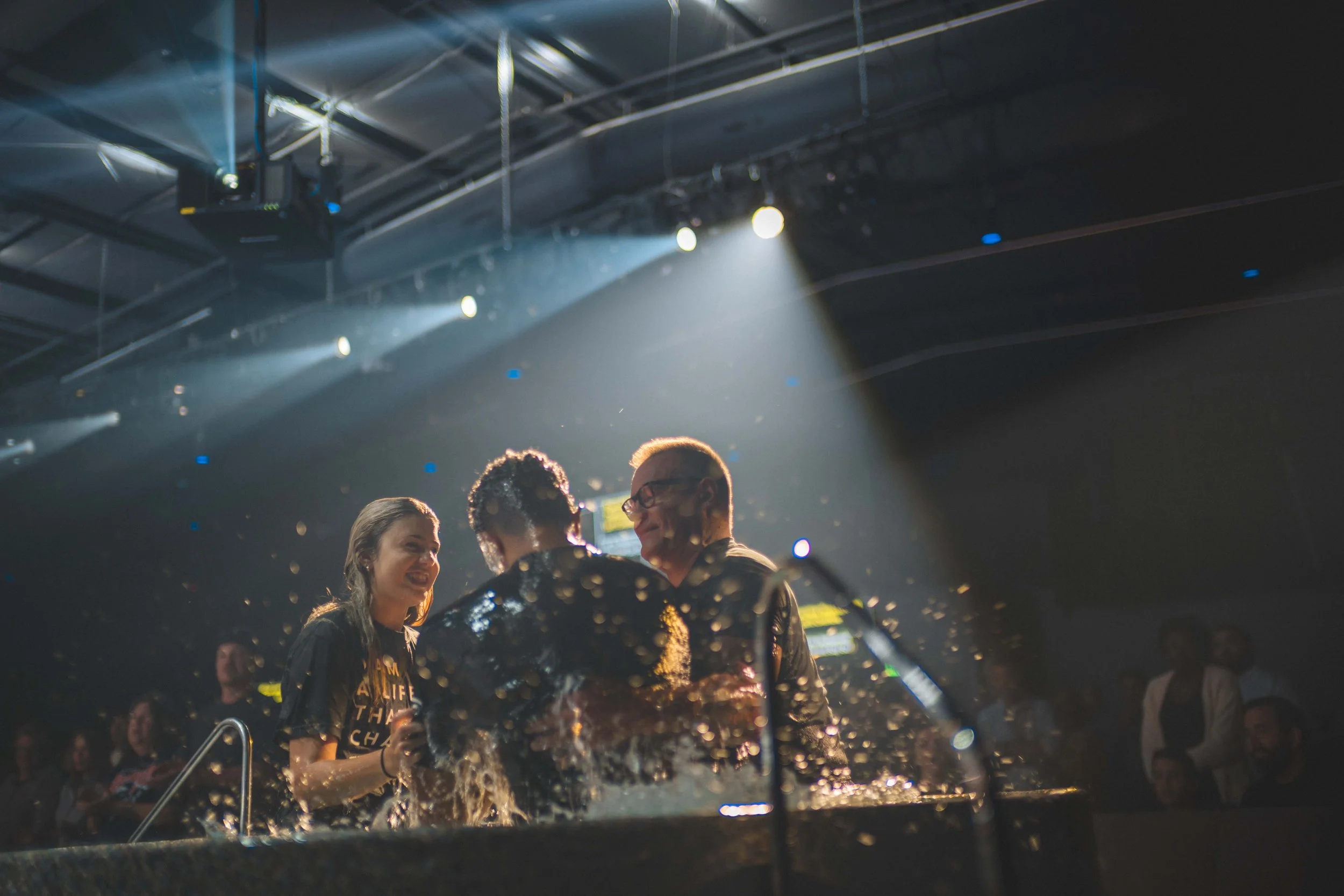 People laughing and smiling in a dunk tank at a party or event, with water splashing as one person is dunked, under stage lights in a dark indoor setting.