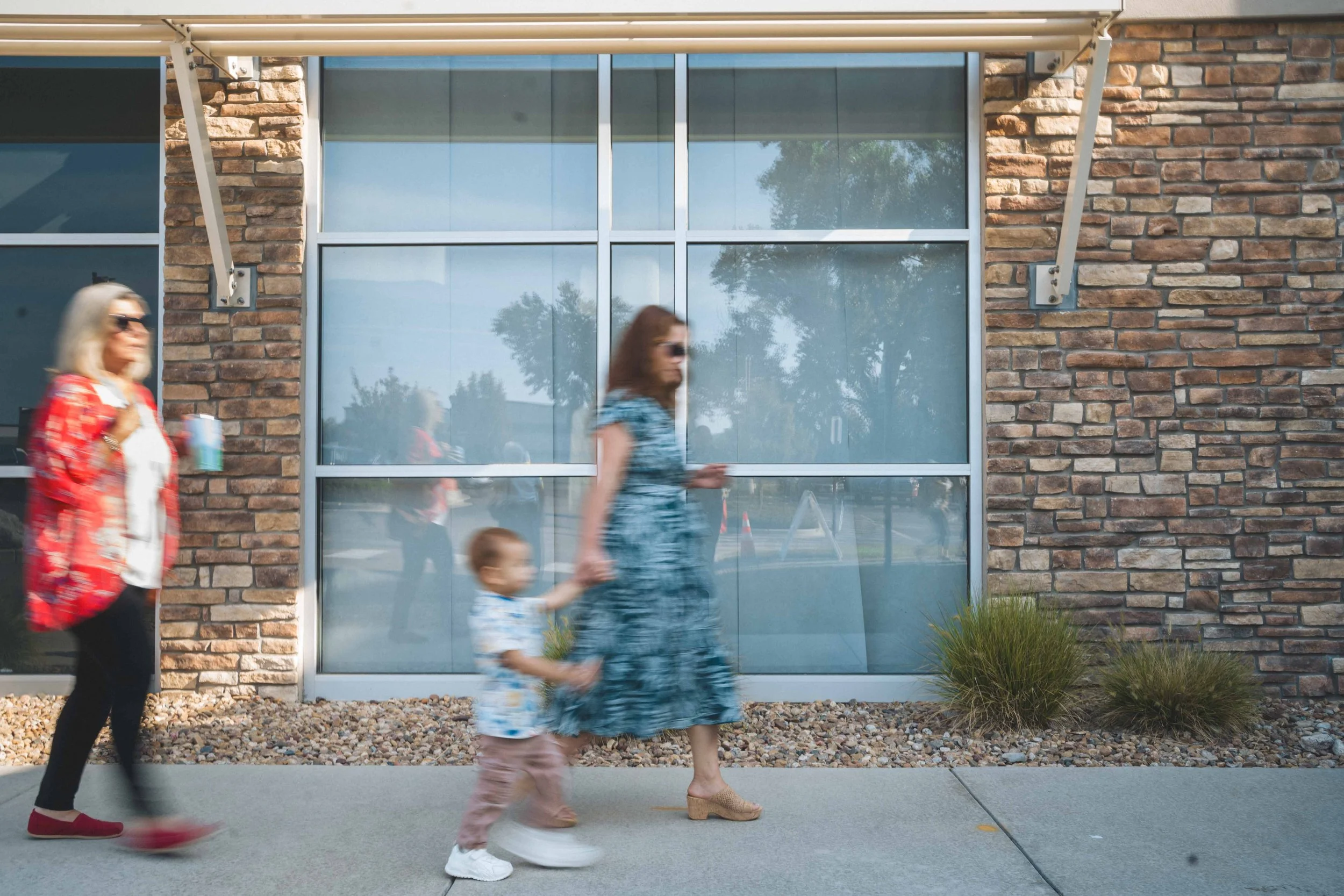 Three people walking in front of a modern brick building with large glass windows. One woman with gray hair and sunglasses holding a drink, a young woman in a patterned blue dress, and a young boy holding the young woman's hand.