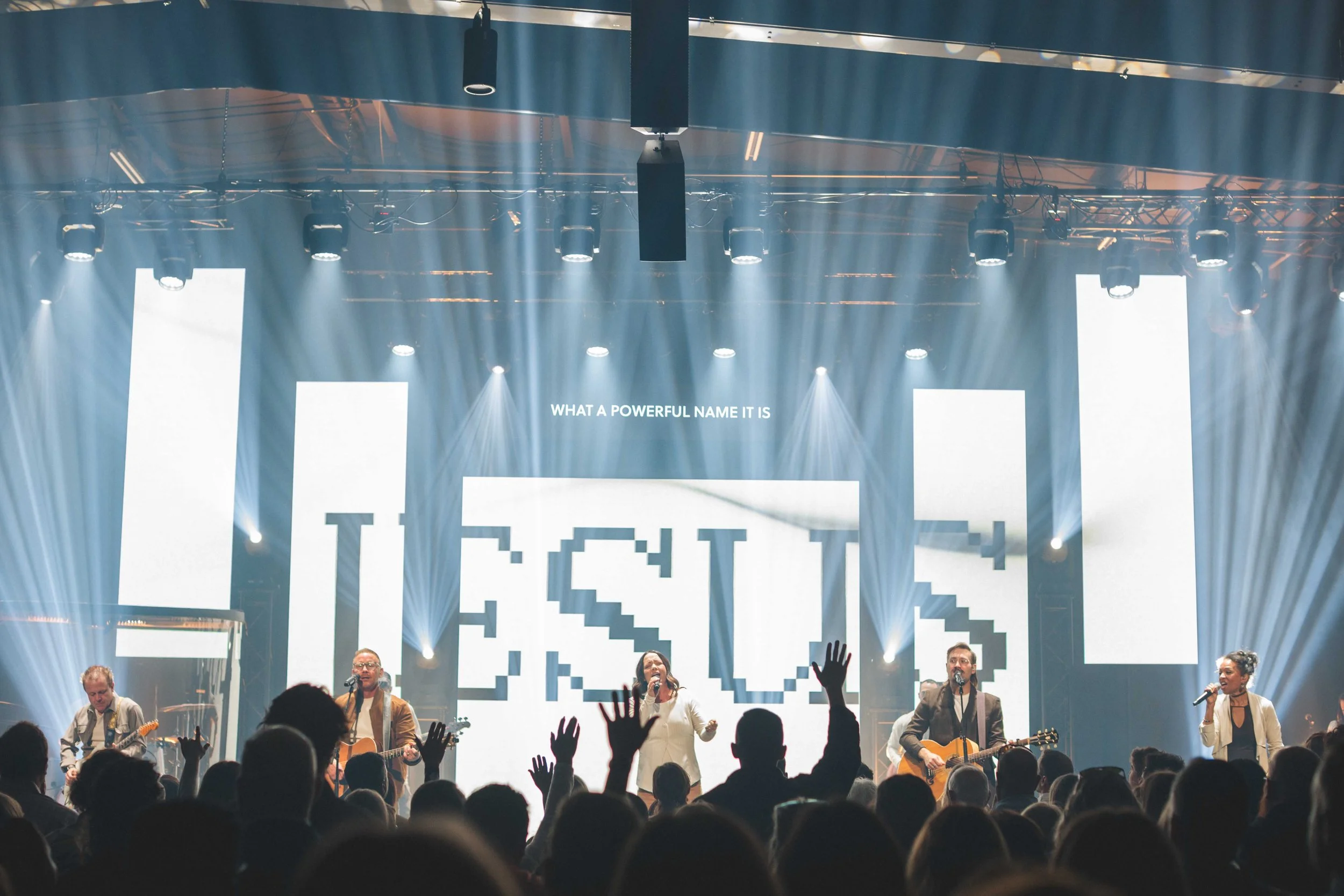 A live concert with a band performing on stage, with five musicians playing guitars and singing. The stage features large LED screens displaying abstract pixelated text and the phrase 'WHAT A POWERFUL NAME IT IS.' The audience is visible in the foreg
