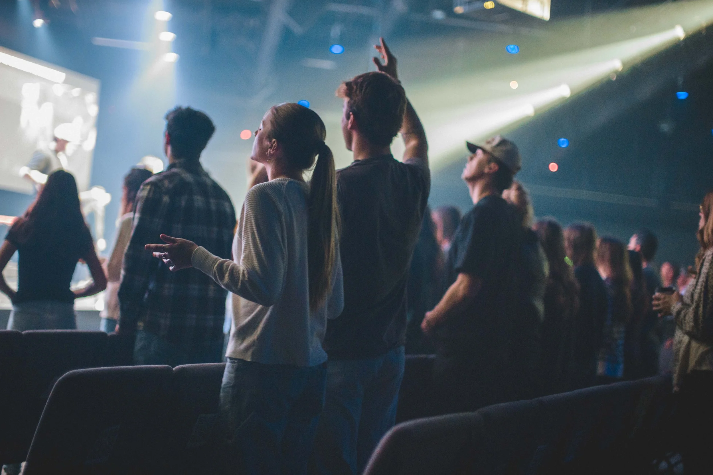 Audience members at a concert or event standing and enjoying the performance, with some raising their hands, in a dimly lit venue with stage lights and a large screen showing a live feed.