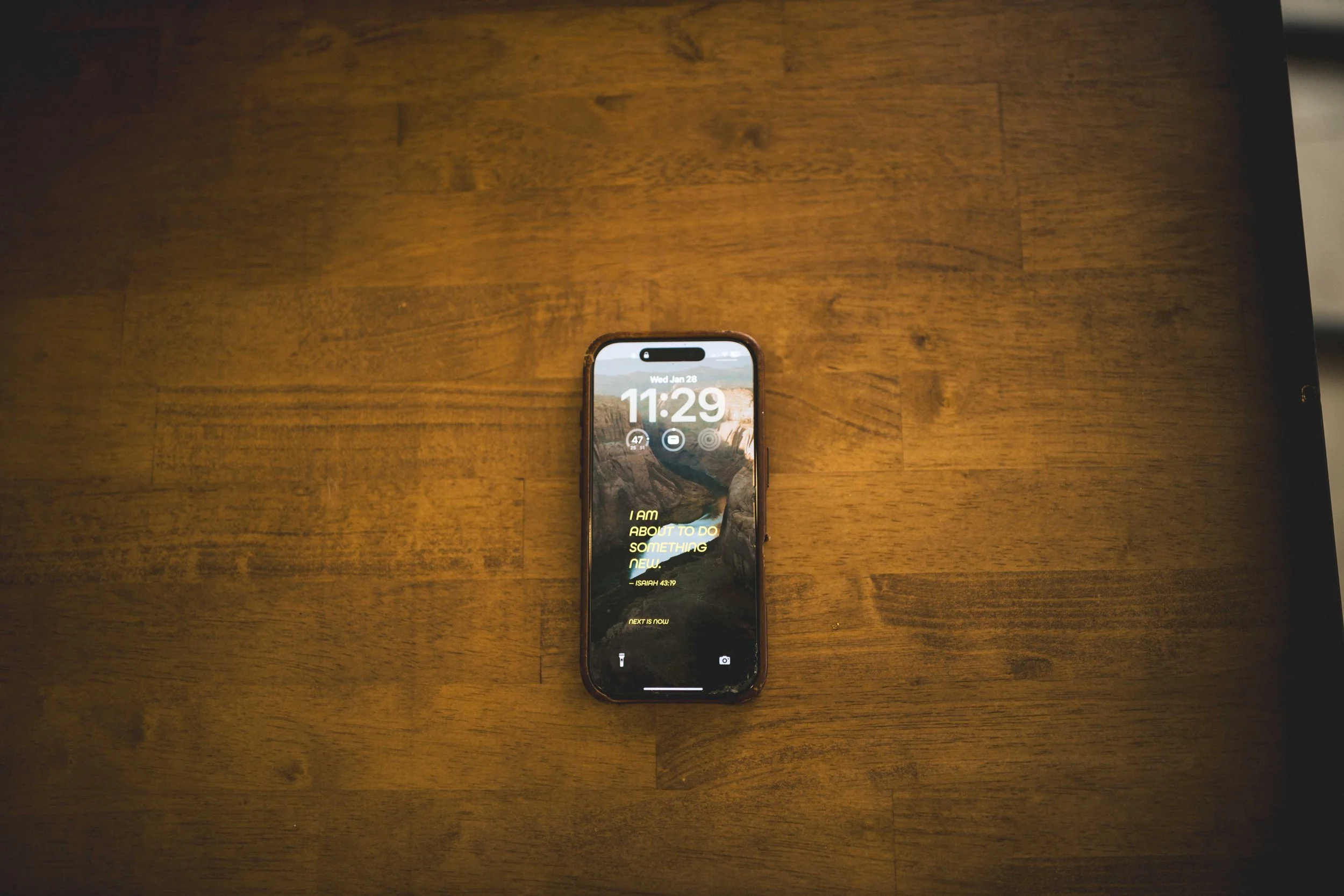 A smartphone with a mountain landscape wallpaper, displaying the time 11:29 and date Wednesday, January 28, lying on a wooden table.