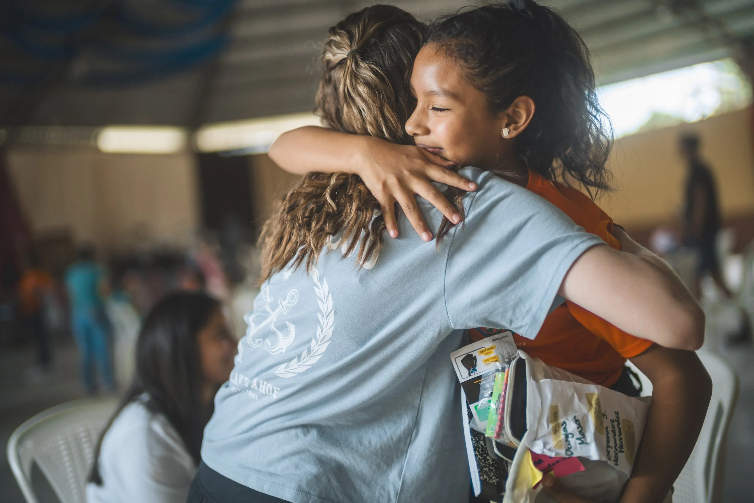 Two women hugging each other warmly in an indoor setting, with other people in the background.