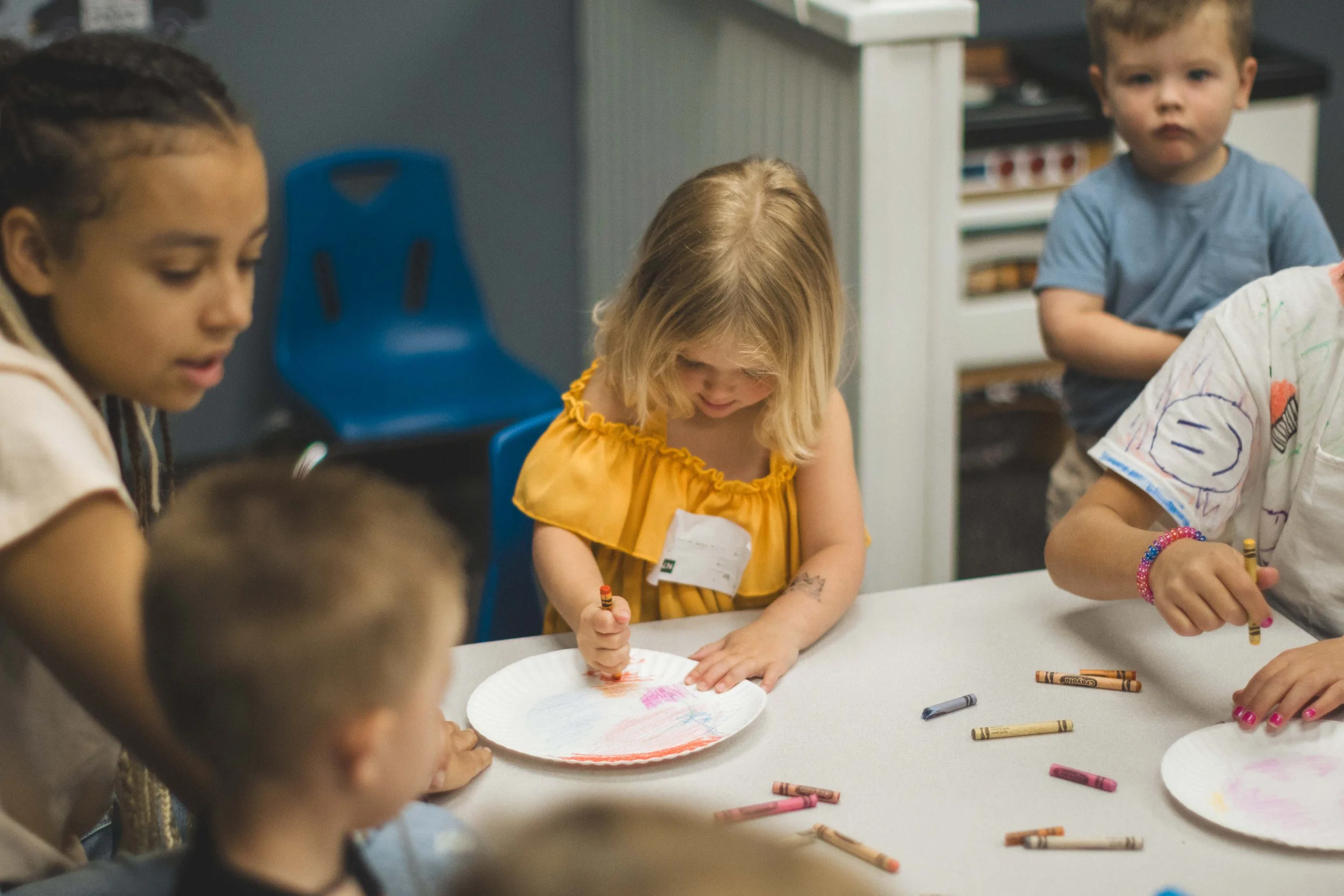 Group of young children sitting around a table, coloring with crayons on paper plates.