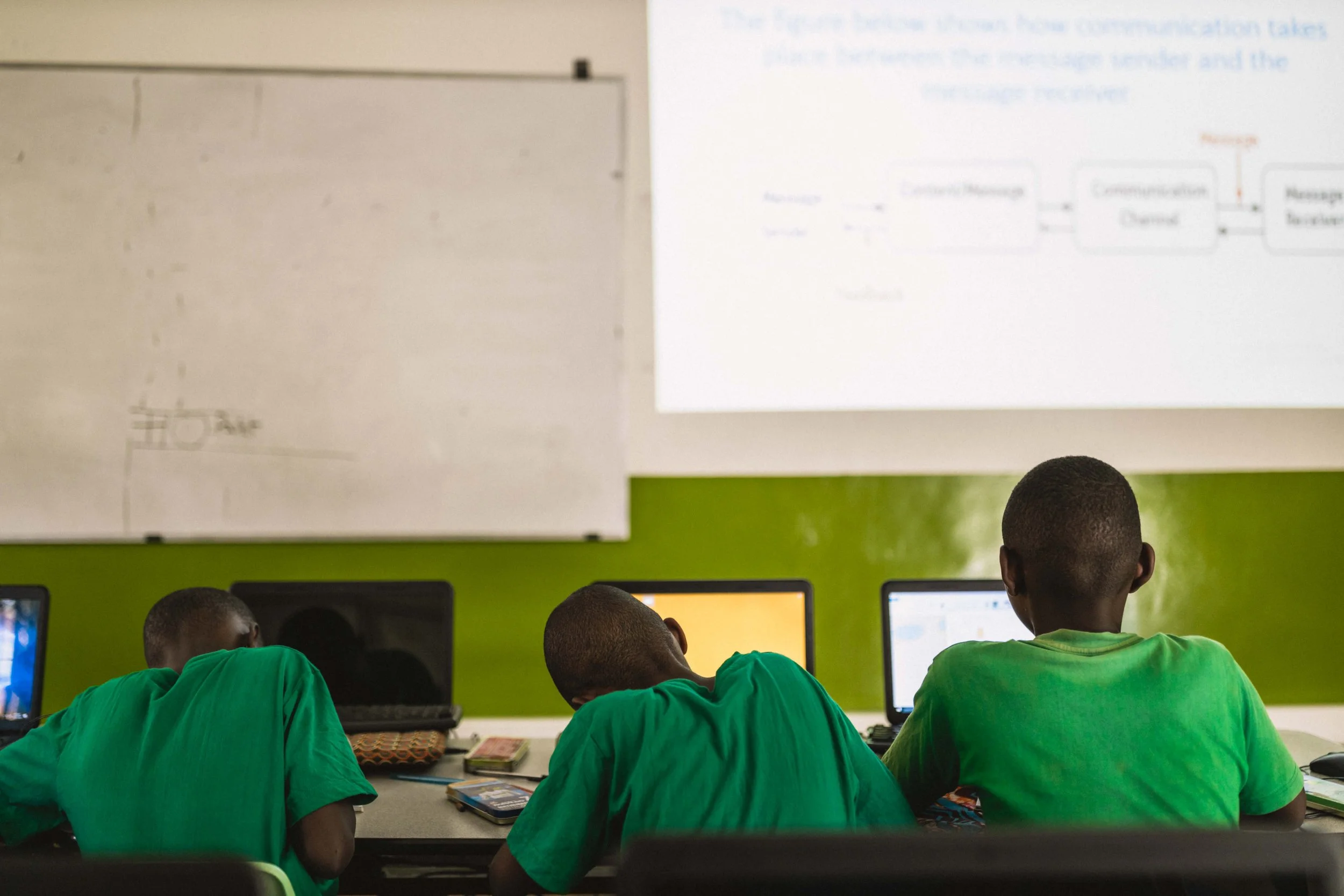 Three students in green shirts sit at desks with computers in a classroom, facing a large projection screen displaying a presentation.