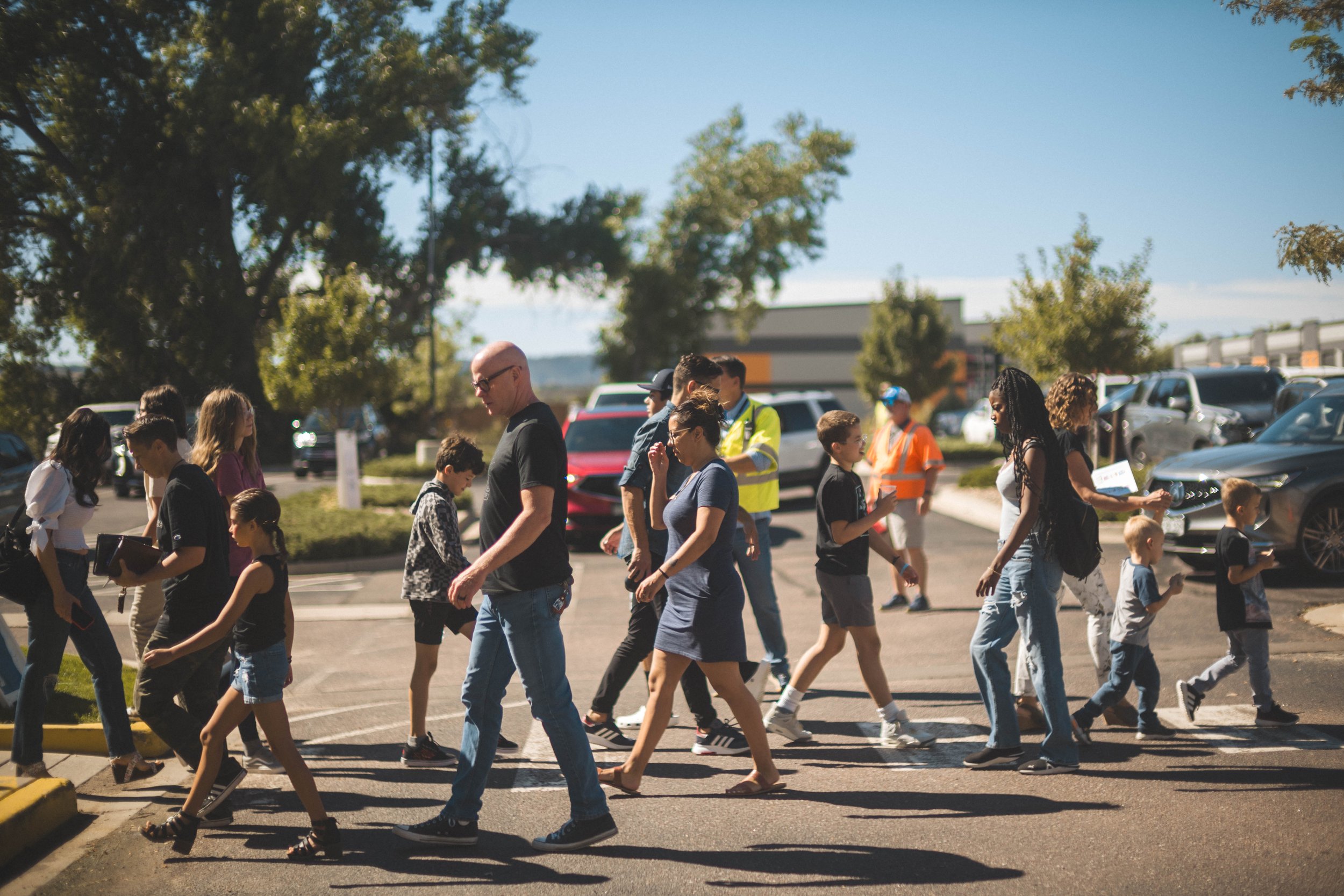 Group of people crossing a parking lot, including children and adults, some wearing casual clothes and safety vests, on a sunny day.