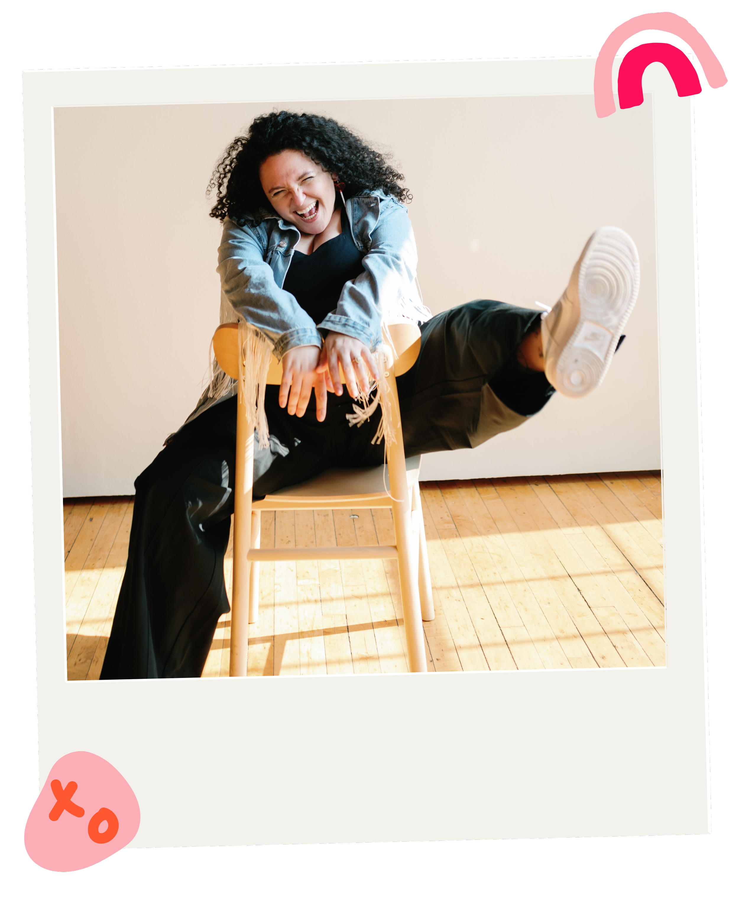 A woman laughing joyfully while sitting on a chair with one leg raised and holding a table, with a woman lying across her lap, in a room with wooden flooring.