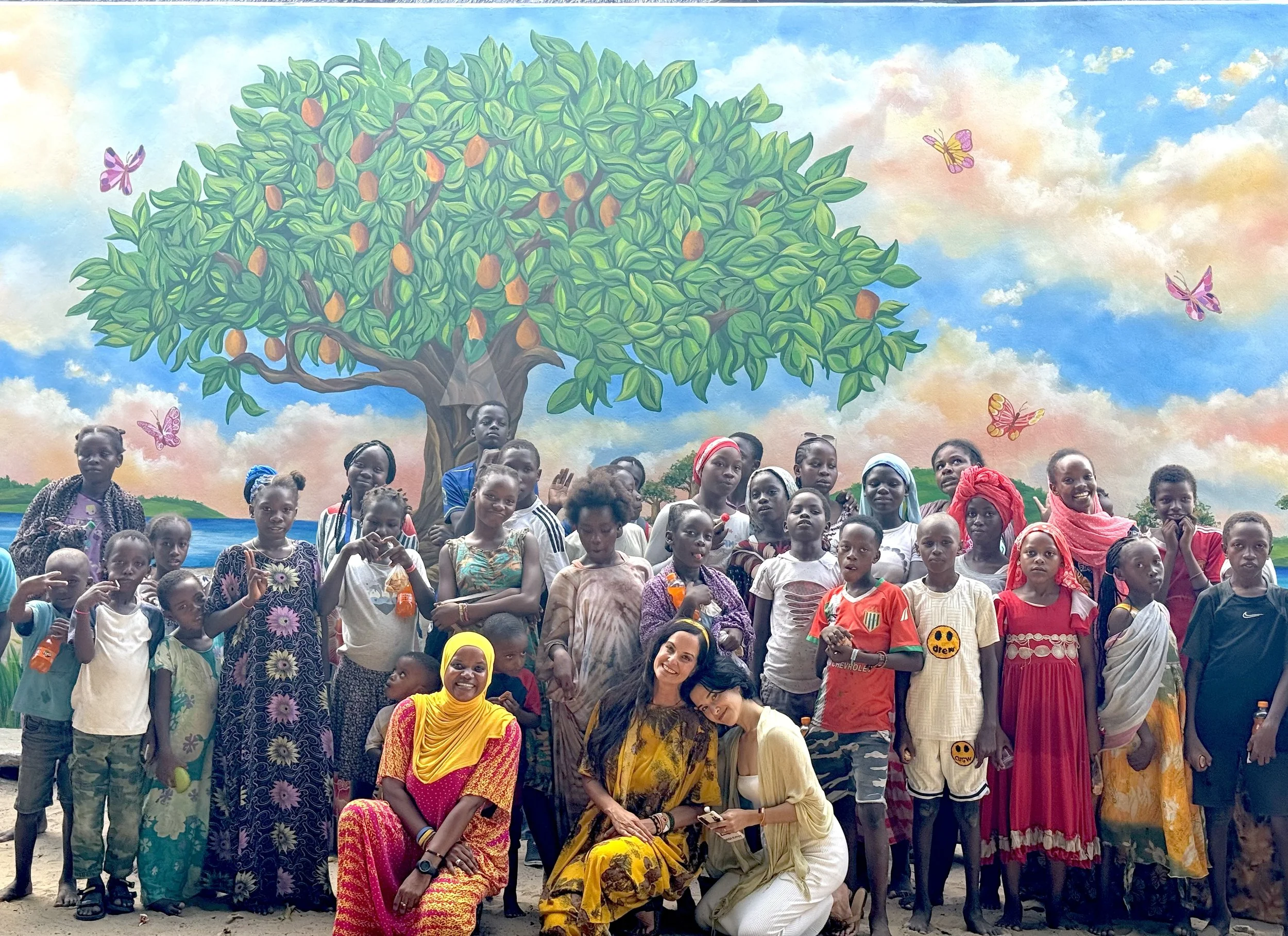 Group of children and women posing in front of a colorful mural with a tree, butterflies, sky, and water scene.