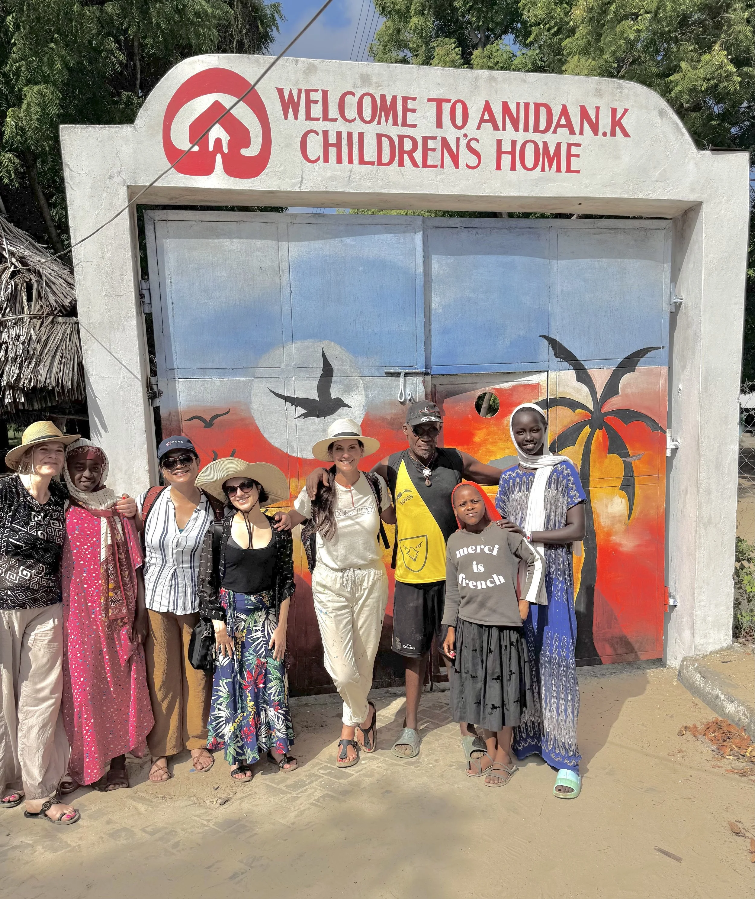 Group of people standing in front of a colorful painted gate with the sign 'Welcome to Anidan.K Children's Home' above it.