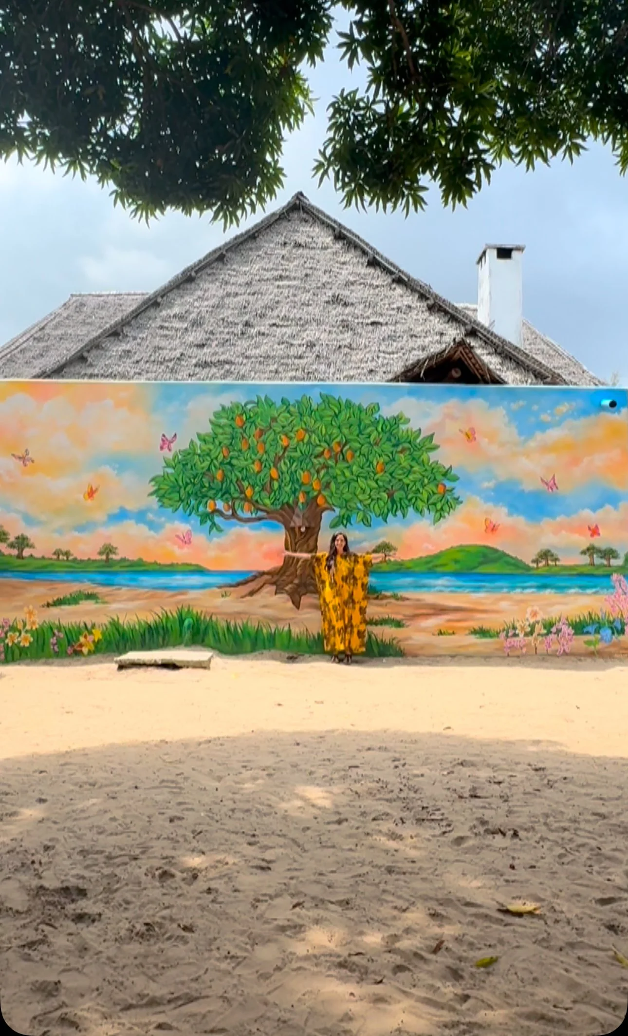 A mural of a large green tree with orange fruit, butterflies, and a woman in a yellow dress standing in front of it, painted on a wall at a scenic outdoor location with a sandy ground and sky in the background.