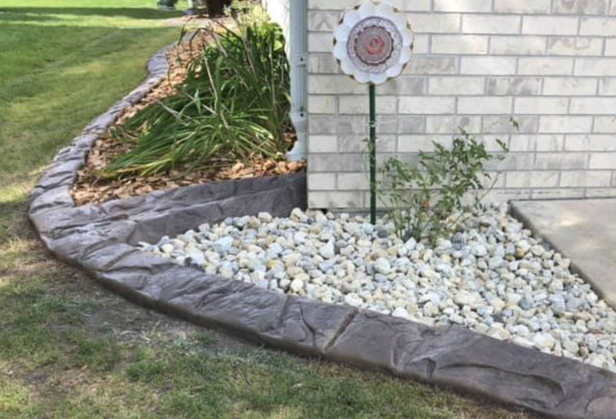 A landscaped yard with a flower bed edged with a faux stone border, filled with white rocks and some plants. There is a decorative flower stake and a brick house in the background.