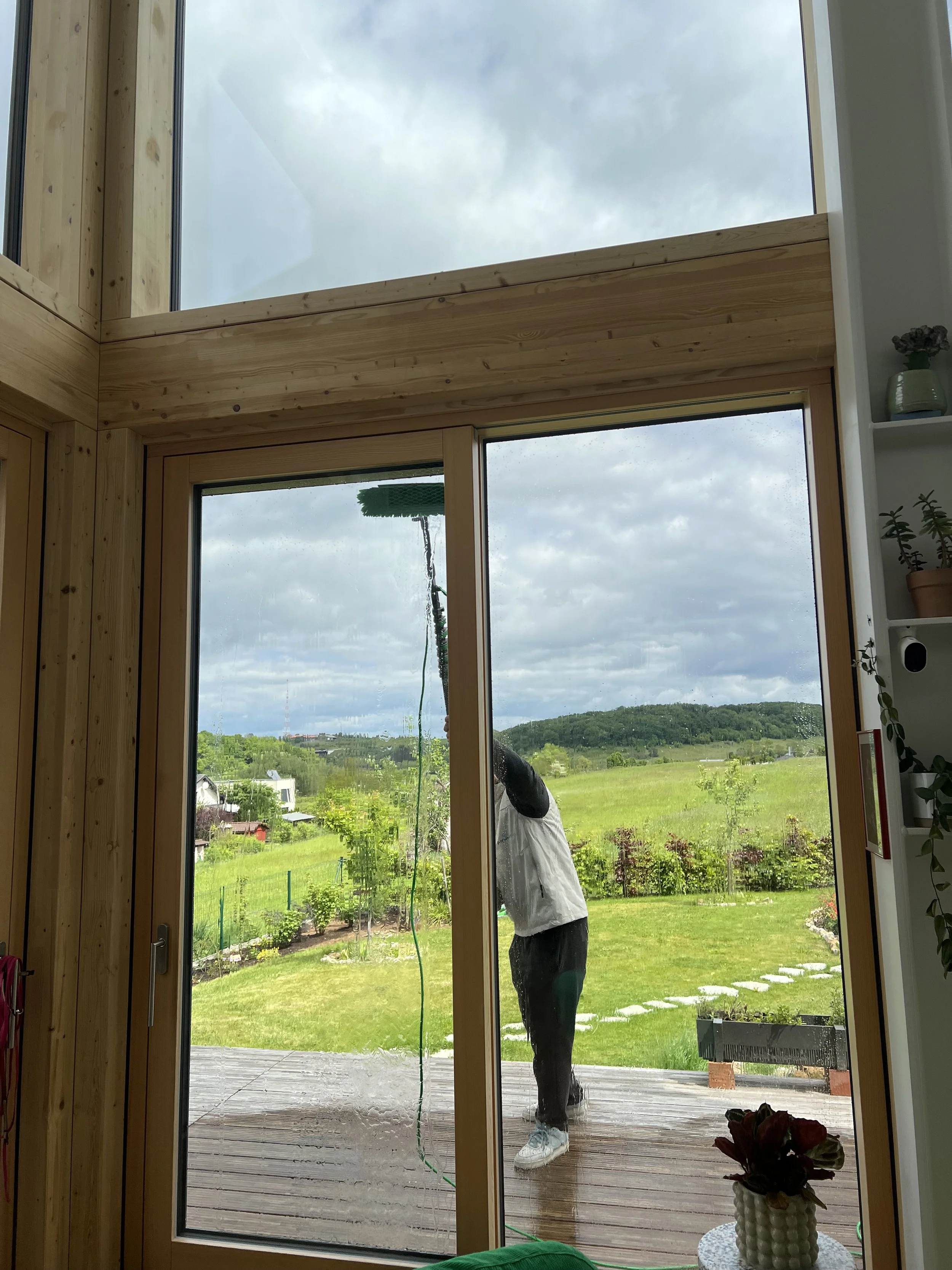 Une personne nettoie une grande baie vitrée dans un jardin verdoyant, avec des nuages dans le ciel, vue depuis l'intérieur d'une maison en bois.