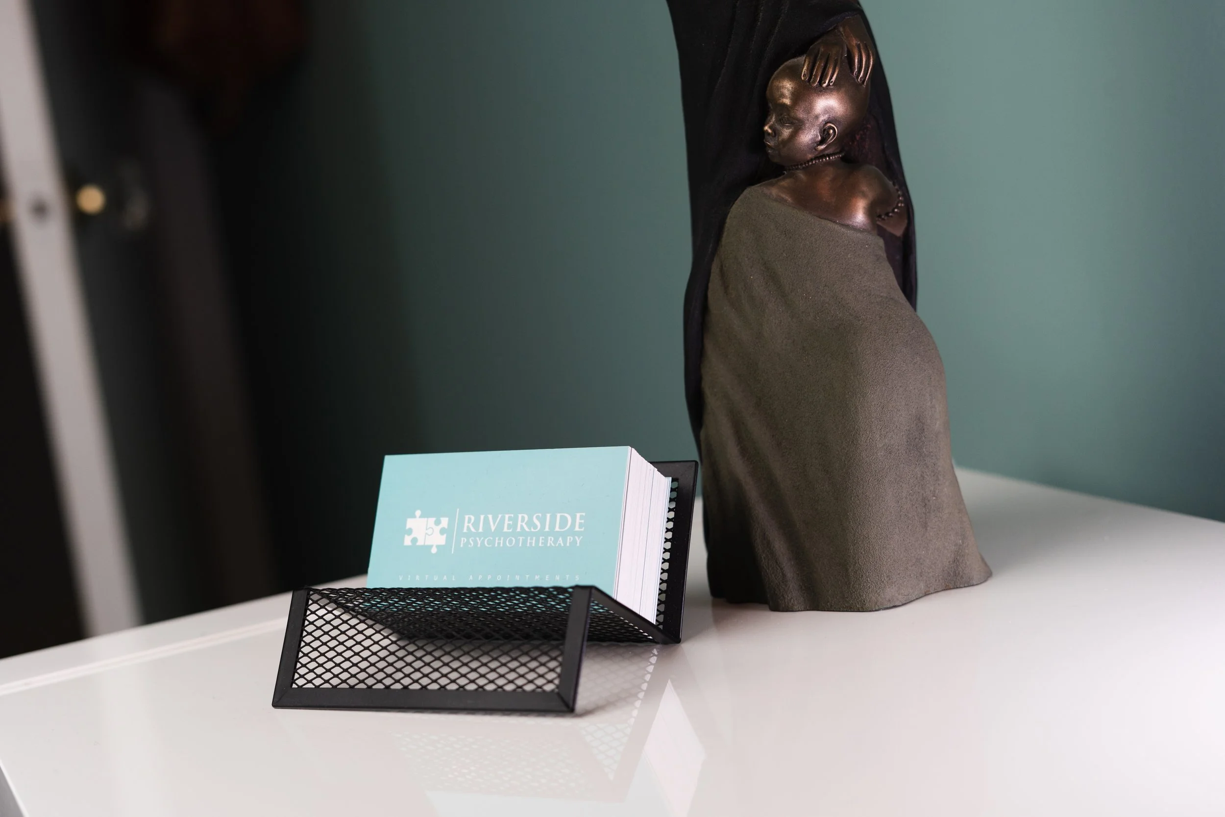 A black metal mesh business card holder on a white table with a blue card that reads 'Riverside Psychotherapy' next to a decorative sculpture of a person with a headscarf and a child on their shoulder.