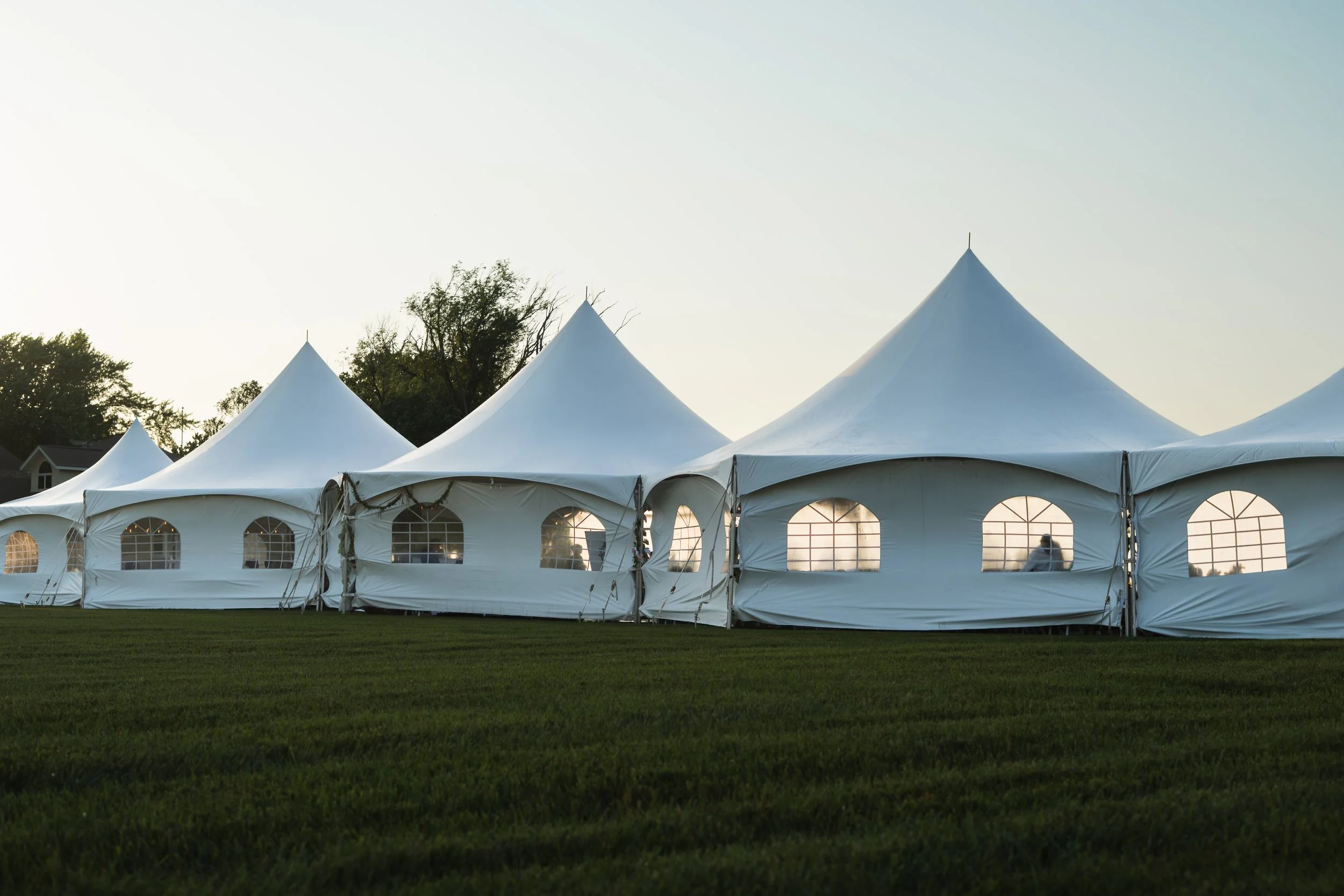 A row of large white event tents with pointed tops and arched windows, set on a grassy field during sunset.