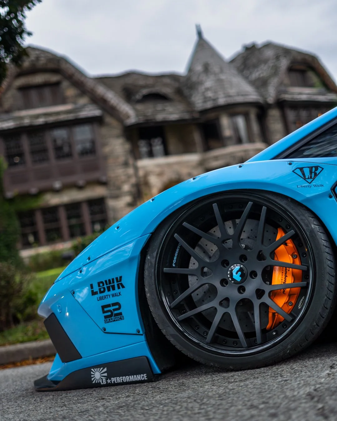Close-up of a blue race car with orange brake calipers and black wheels, parked in front of a large stone and wood house.