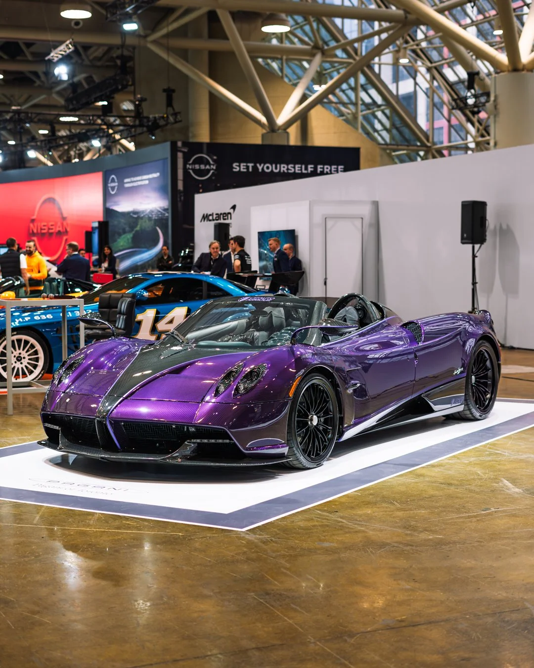 A purple sports car on display at an indoor car exhibition, with other race cars and automotive brand banners in the background.