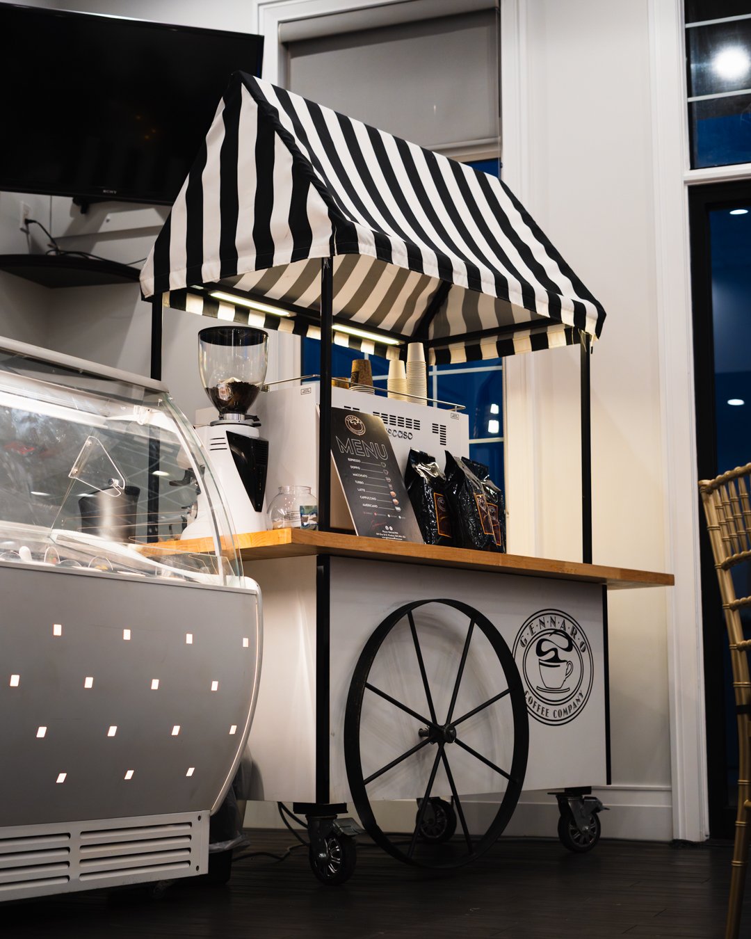 A coffee stand with a black and white striped canopy, a menu board, coffee bags, a coffee grinder, and a logo on the side of the cart that reads 'Fennaro Coffee Company.'