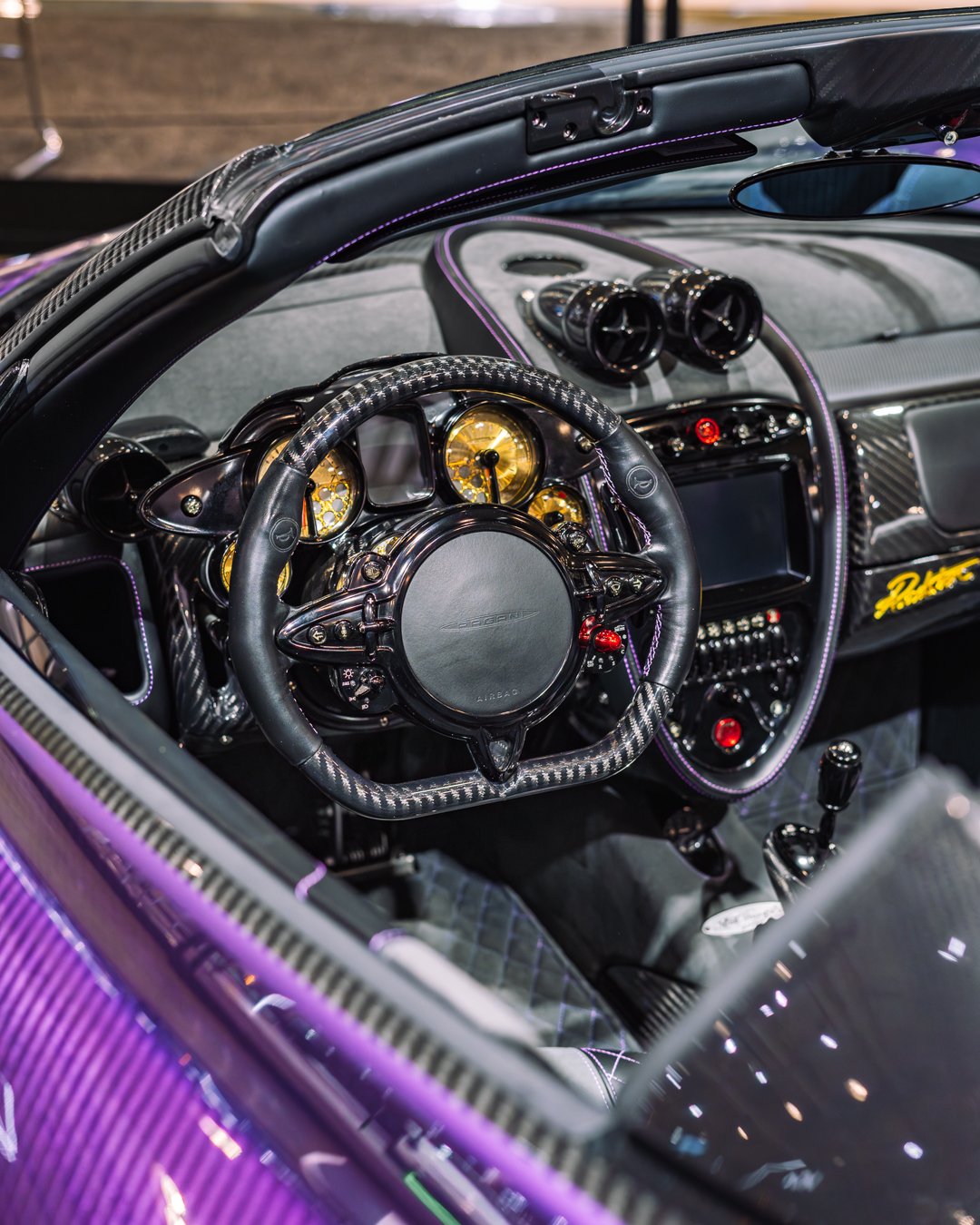 Close-up of a luxury car interior featuring a carbon fiber steering wheel with a central hub marked 'Aston Martin,' golden gauges, black dashboard with purple stitching, and various control buttons.