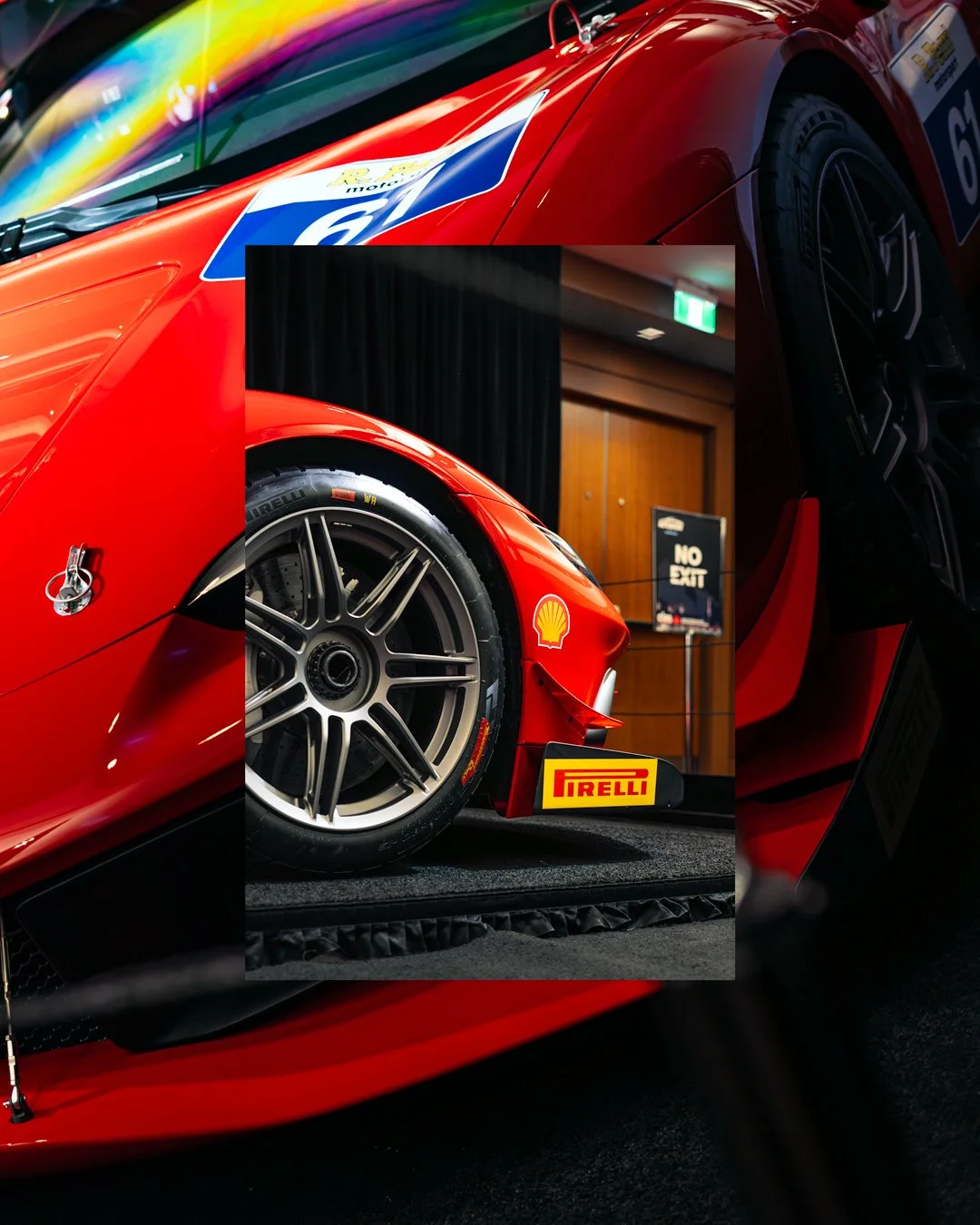 Close-up of a red race car with Pirelli tires, Shell logo, and Pirelli signage at an indoor exhibition, with a background showing a 'No Exit' sign and wooden doors.