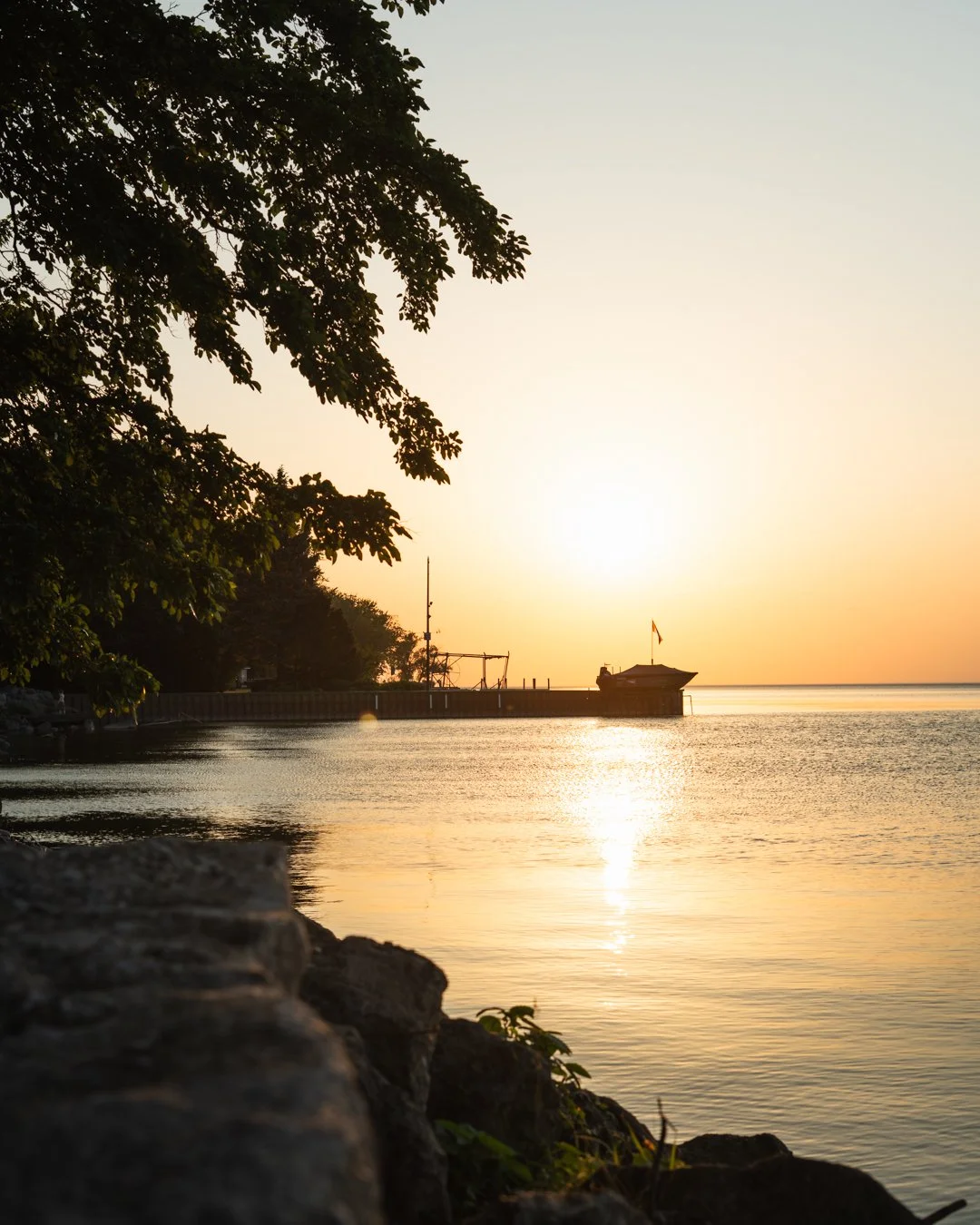 Sunset over a calm body of water with a small pier or dock, trees on the left, rocks in the foreground, and a boat with a flag on the dock.
