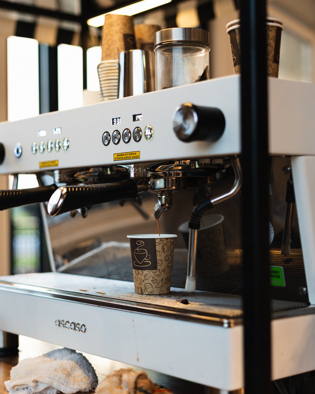 A white espresso machine brewing coffee into a paper cup in a coffee shop. Several disposable cups are on top of the machine.