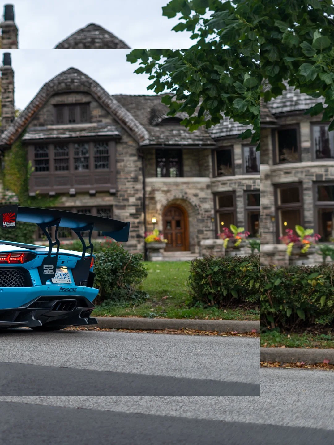 A blue sports car with a large rear wing parked in front of a large, stone house with multiple windows and a wooden front door, partially obscured by a tree and shrubs.