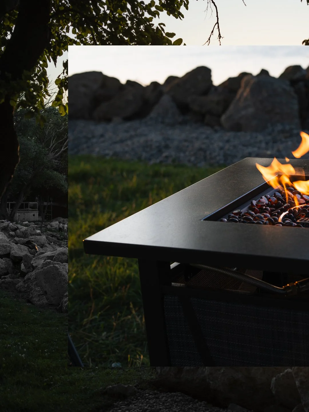 A metal fire pit with a small flame burning among stones, set outdoors on grass during dusk or sunset.