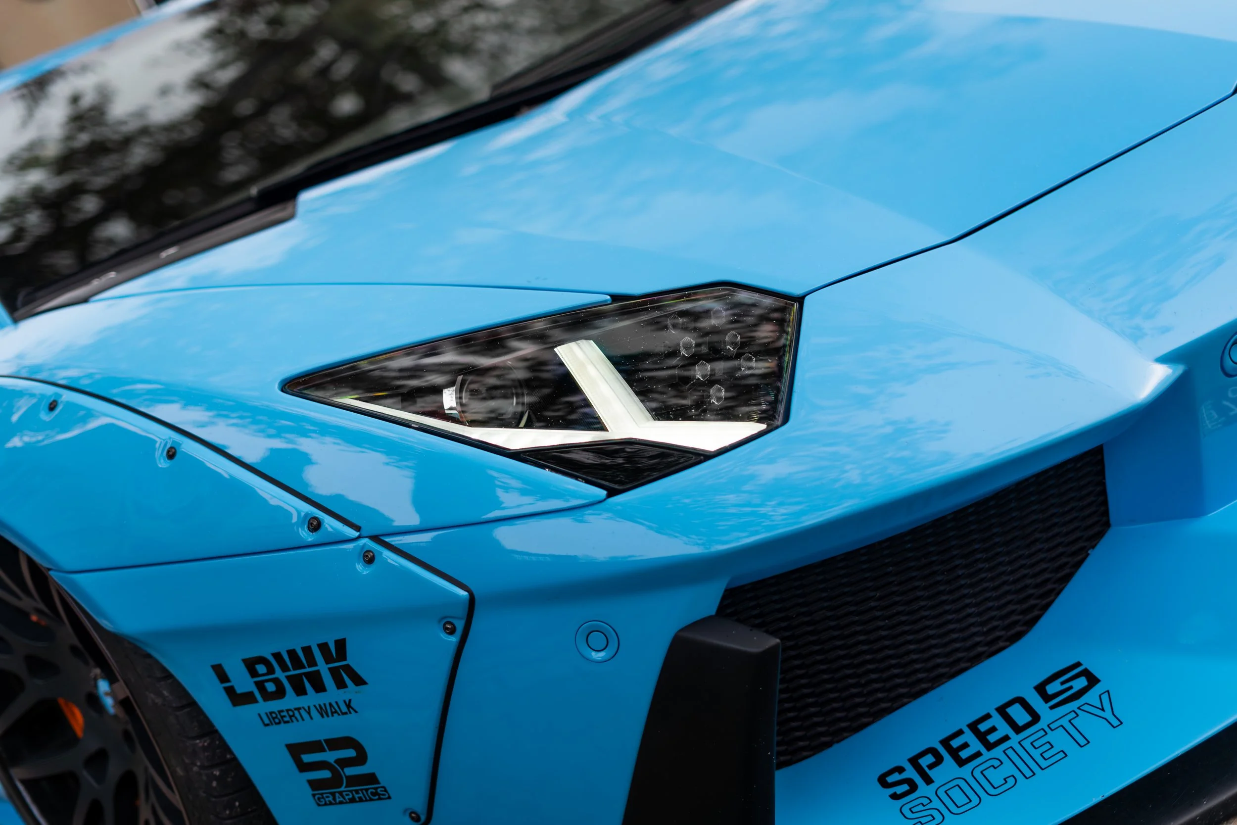 Close-up of a blue sports car's front end, showing the headlight, fender, and decals including 'Liberty Walk' and 'Speed Society'.
