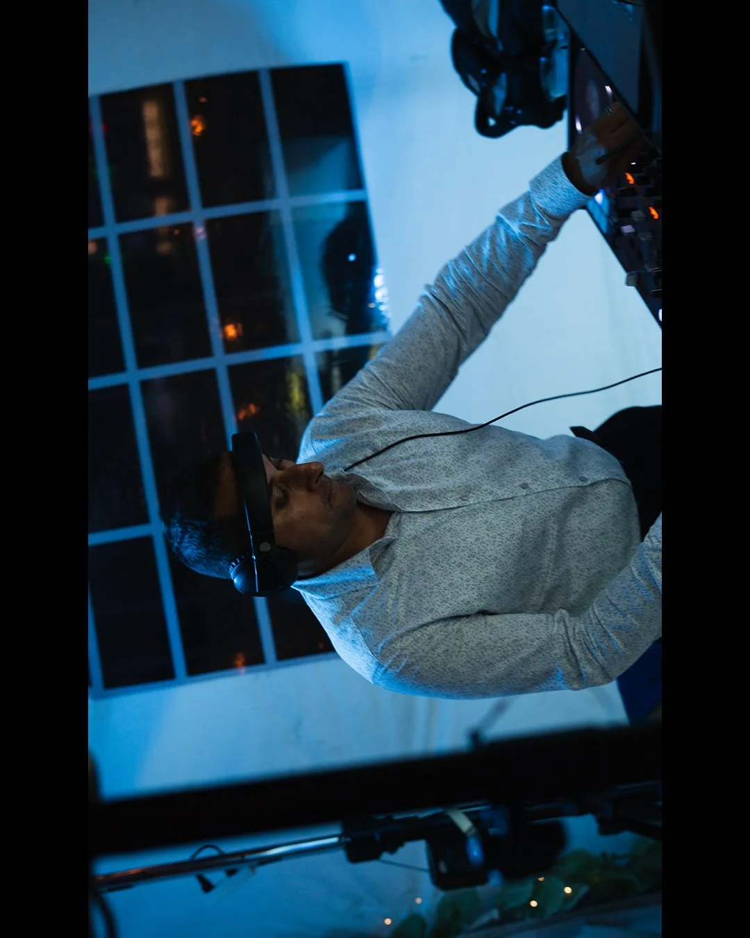 A DJ wearing headphones and a gray sweater is playing music on a DJ controller at night. A large window behind him reveals city lights outside.