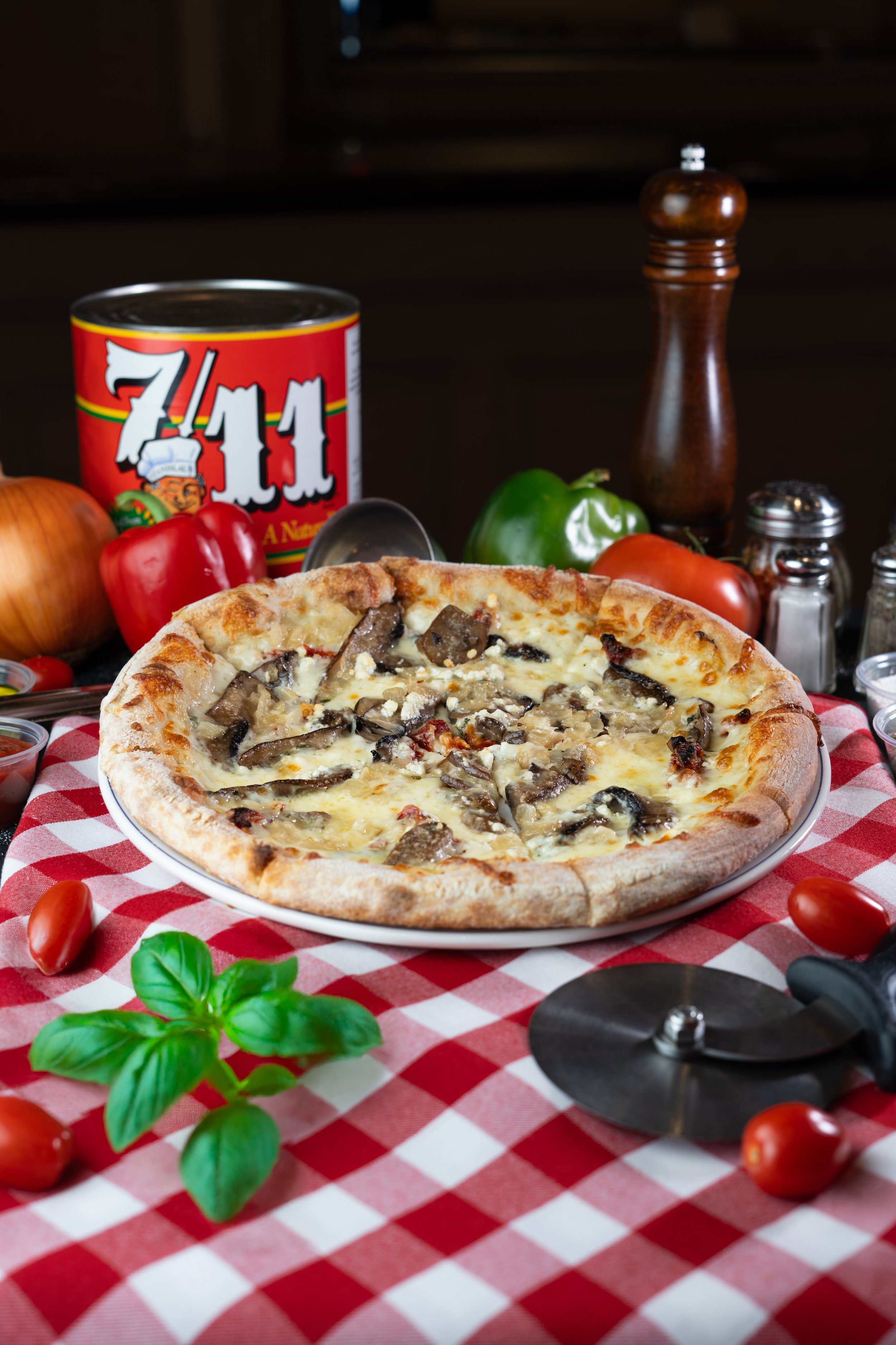 Fresh mushroom and cheese pizza on a white plate, placed on a red and white checkered tablecloth with cherry tomatoes, basil leaves, and pizza cutter nearby, with canned food and fresh vegetables in the background.