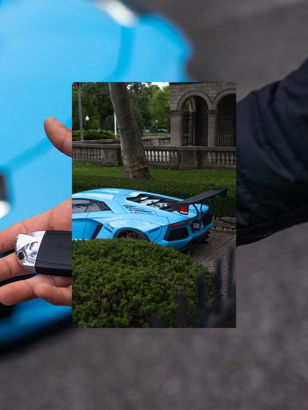 Person holding a Lamborghini key fob in front of a blue Lamborghini sports car parked outdoors near a park and historic building with arches.