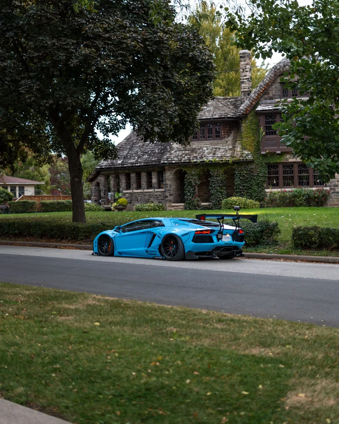 A bright blue sports car with a large rear wing and aggressive aerodynamics parked on a suburban street in front of a large stone house with trees and grass.