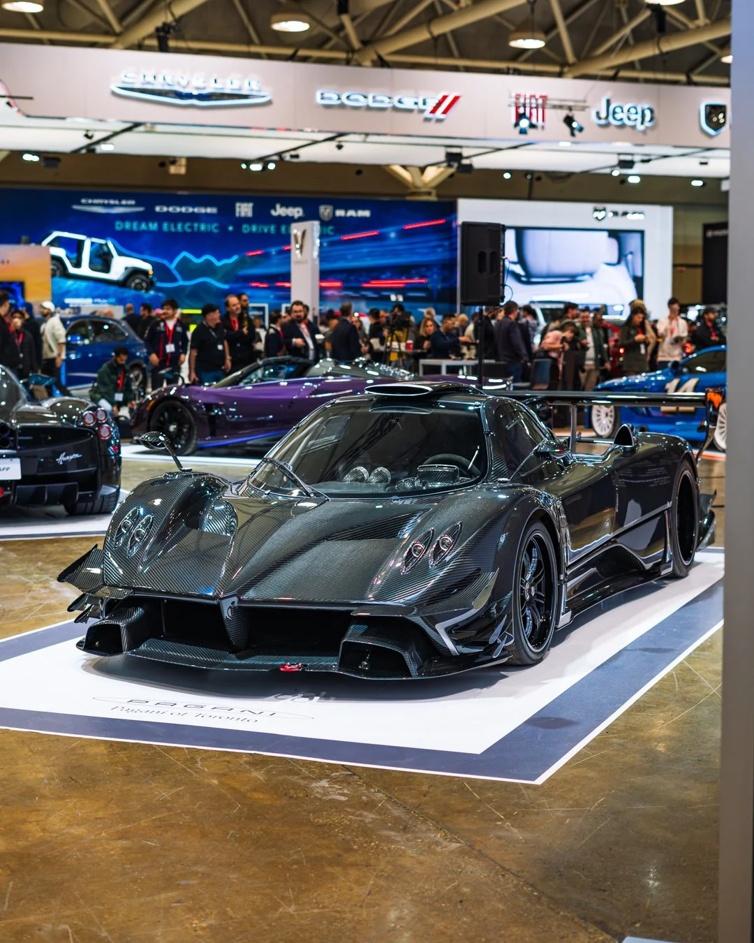 A black carbon fiber racing car displayed at an auto show, with a crowd of people in the background and various car brand logos overhead.